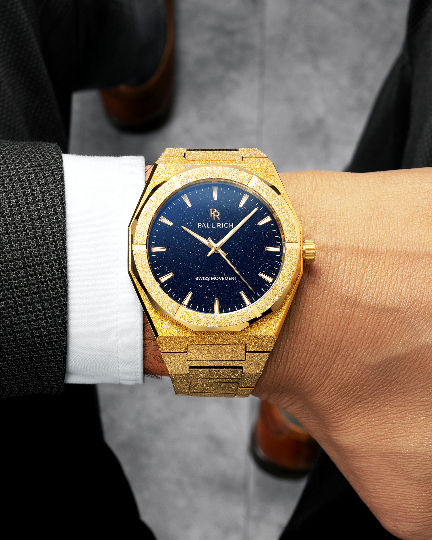 Gold frosted Paul Rich watch with blue aventurine dial worn on wrist, paired with a dark suit, white cuff, and brown shoes.