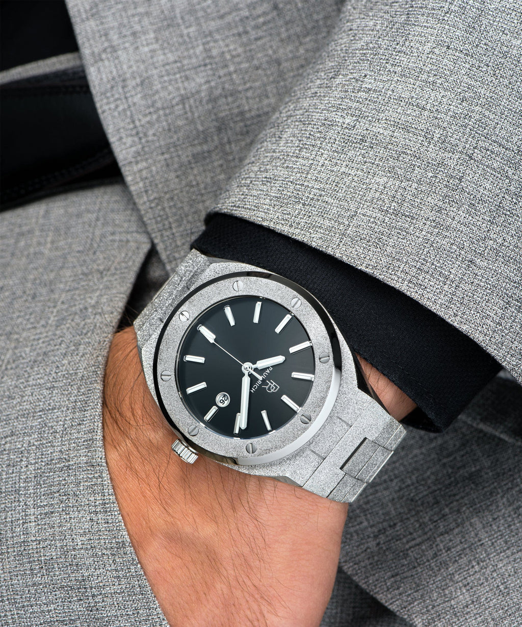Paul Rich watch with frosted silver case and bracelet, metallic black sunray dial, and date window, worn on wrist with grey suit and black cuff.