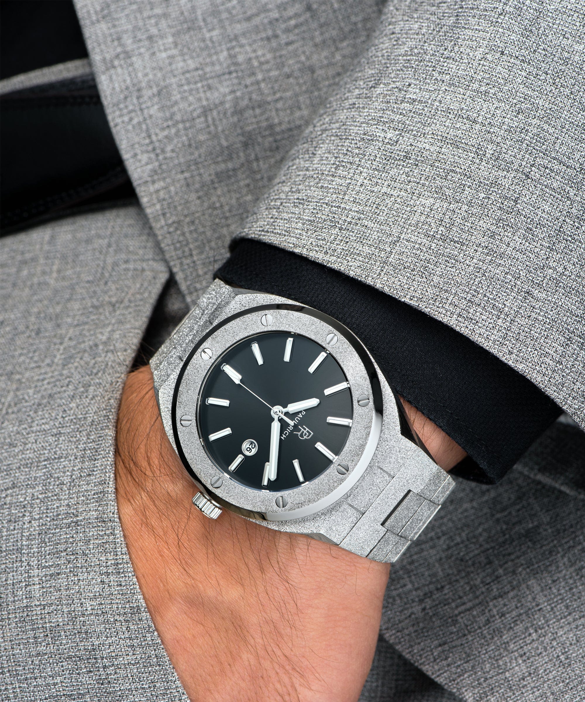Paul Rich watch with frosted silver case and bracelet, metallic black sunray dial, and date window, worn on wrist with grey suit and black cuff.