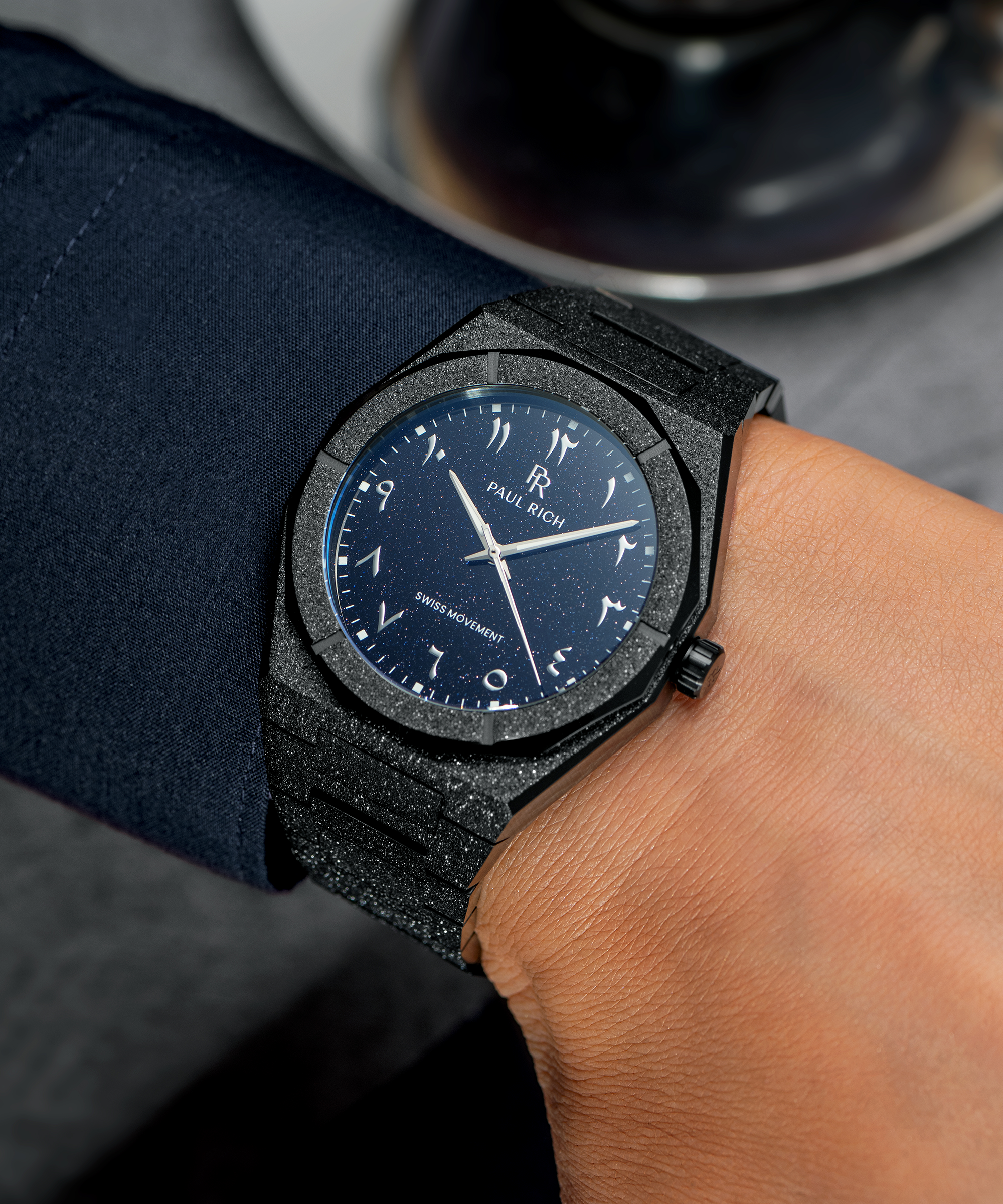 Frosted black watch with a blue aventurine dial and Arabic numerals, worn with a navy cuff, next to a glass cup of espresso.