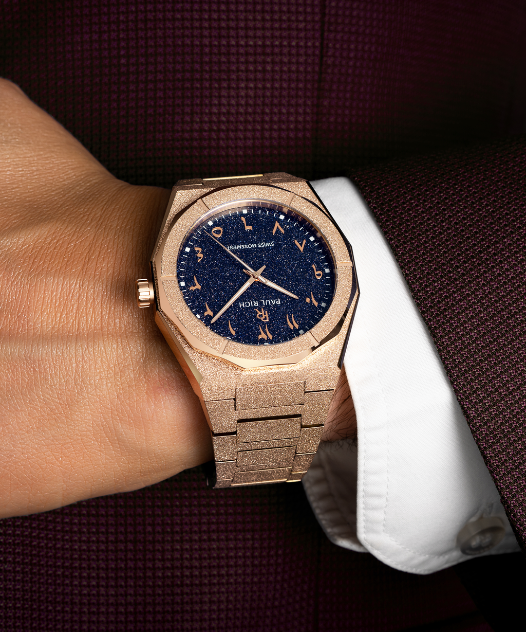 Frosted rose gold watch with blue aventurine dial and Arabic numerals, worn with a white cuff and burgundy textured suit.