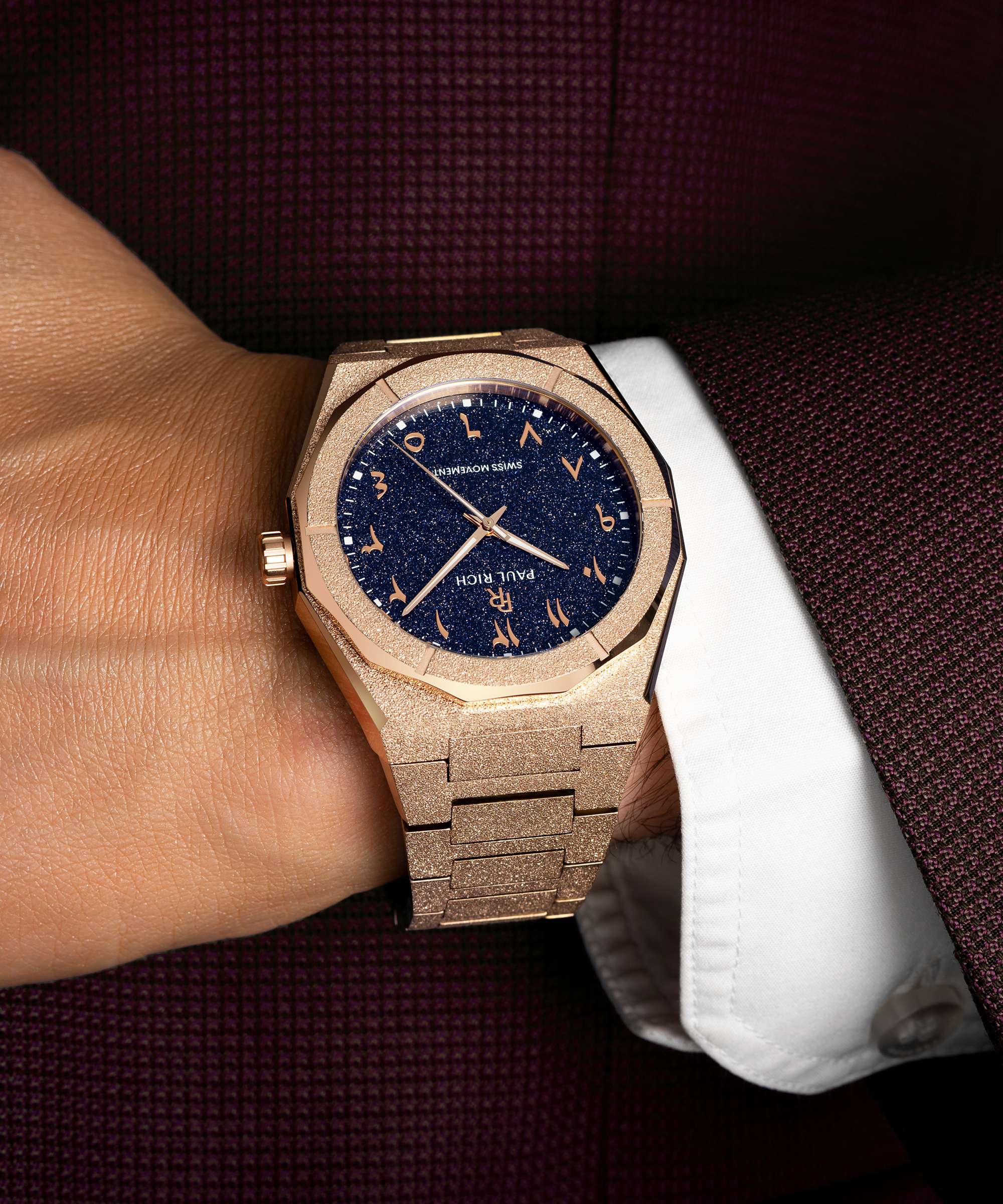 Frosted rose gold watch with blue aventurine dial and Arabic numerals, worn with a white cuff and burgundy textured suit.
