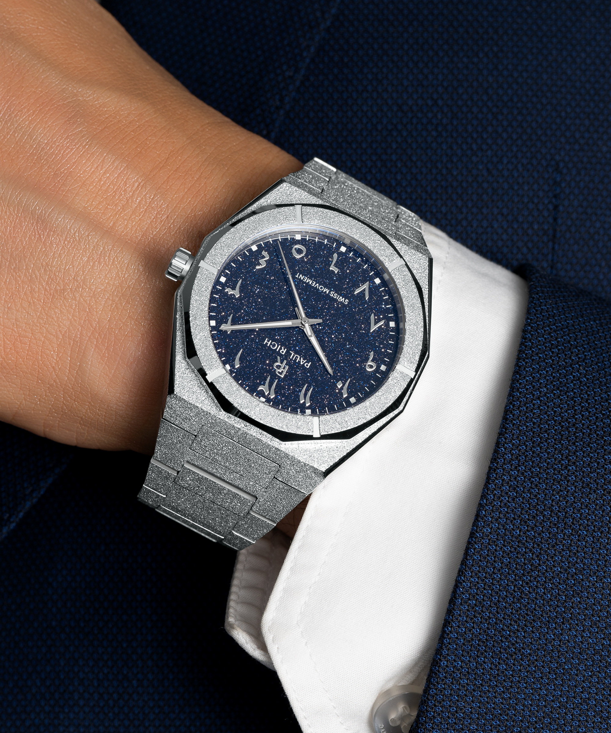 Frosted silver watch with a blue aventurine dial and Arabic numerals, worn with a white cuff and dark blue textured suit.