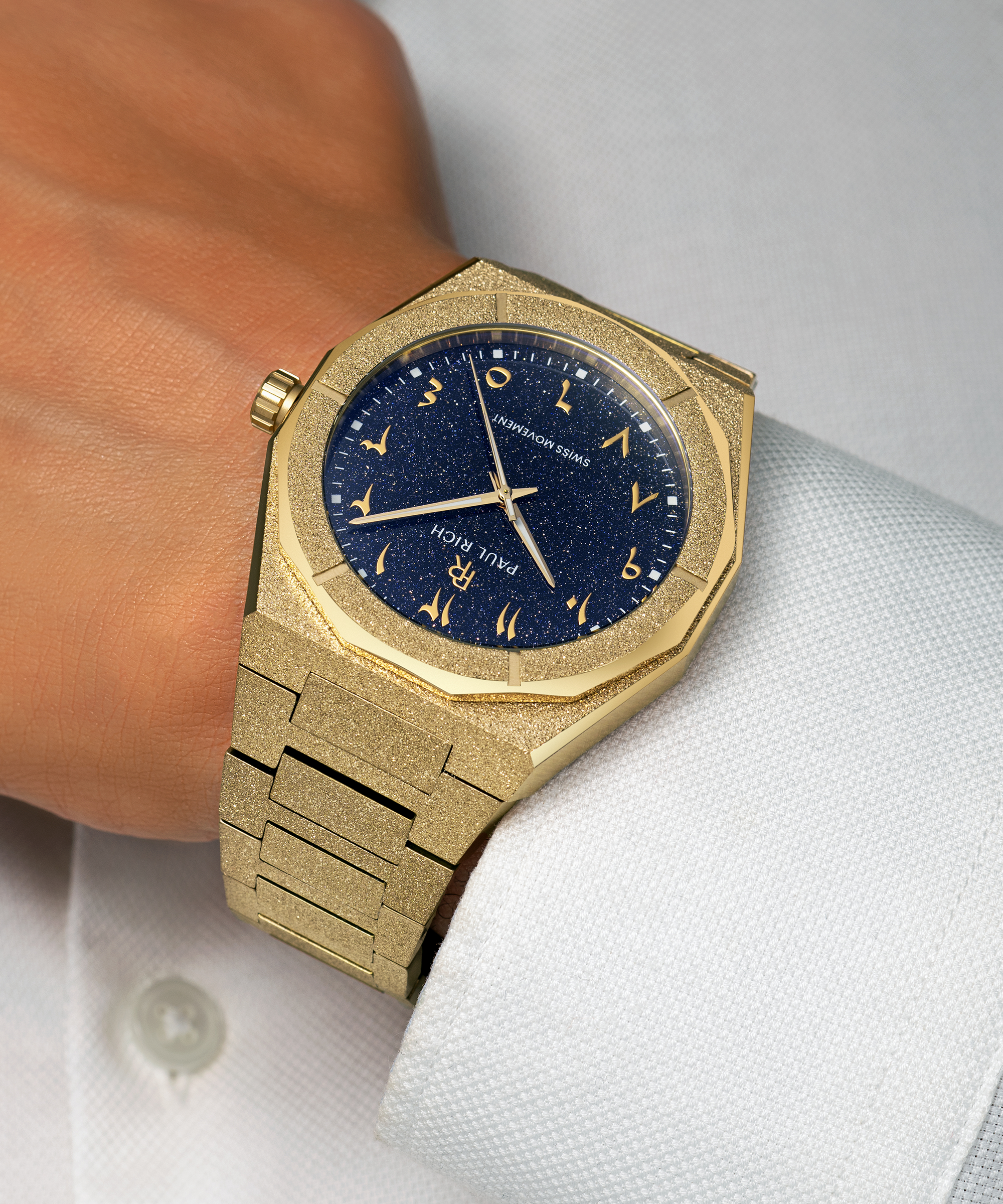 Frosted gold watch with a blue aventurine dial and Arabic numerals, worn with a textured white dress shirt.