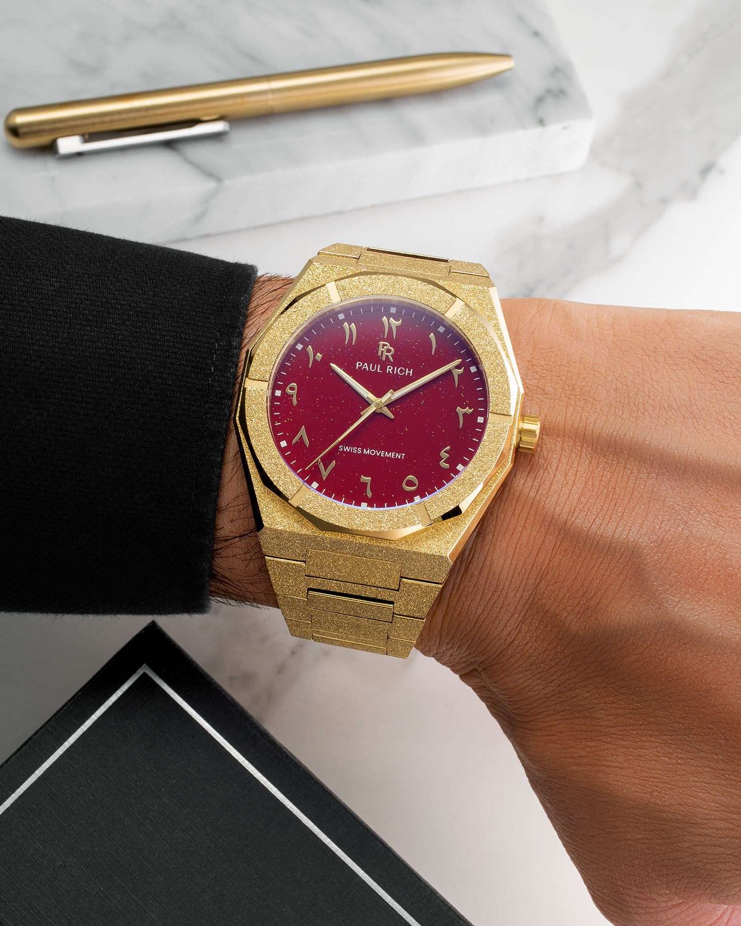 Paul Rich watch with frosted gold case and red aventurine dial with Arabic numerals, worn with a black cuff on a marble desk.