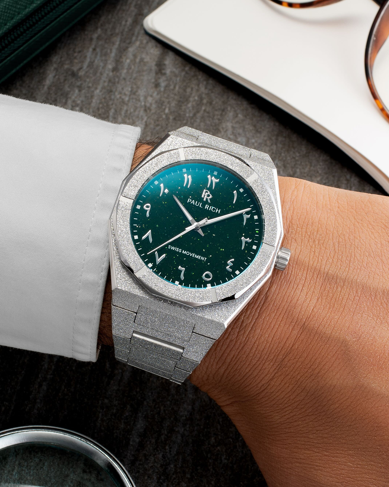 Paul Rich watch with frosted silver case and green aventurine dial with Arabic numerals, worn with a white cuff on a dark tabletop.
