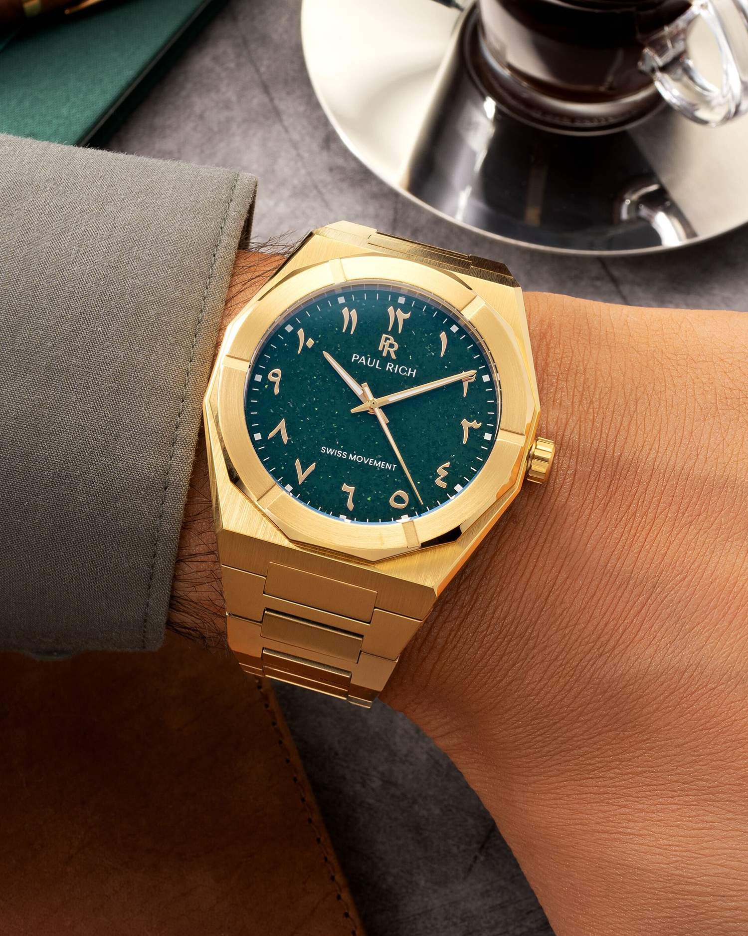 Paul Rich watch with brushed gold case and green dial featuring Arabic numerals, worn with a sage cuff beside a cup of espresso.