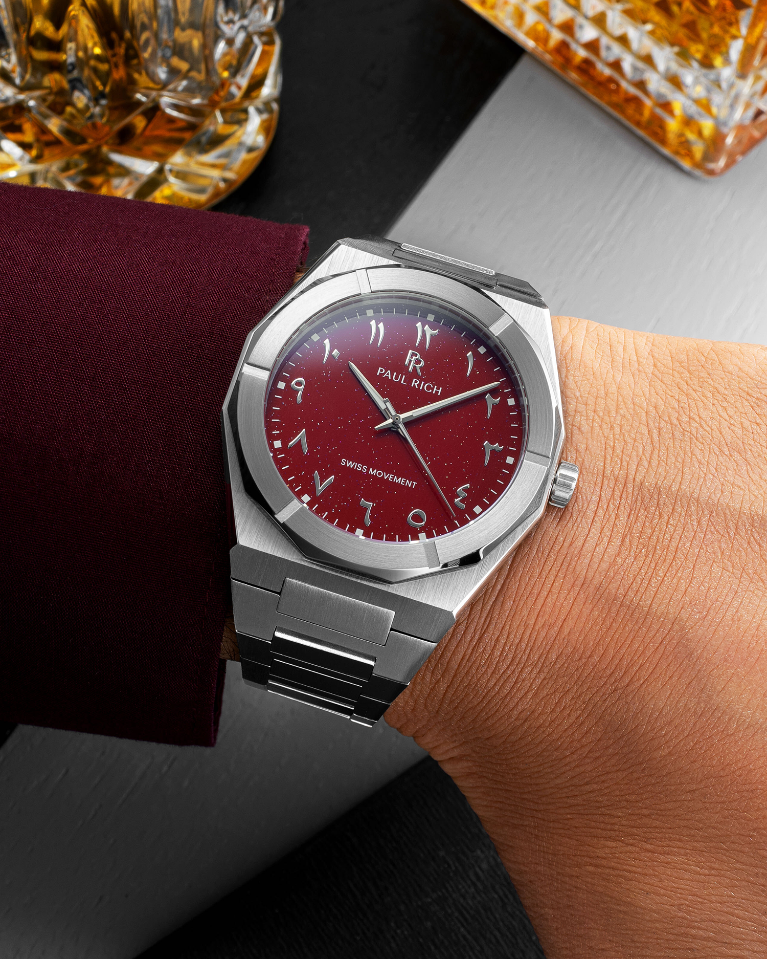 Paul Rich watch with brushed silver case and red dial with Arabic numerals, worn with a burgundy sleeve near crystal whiskey glasses.