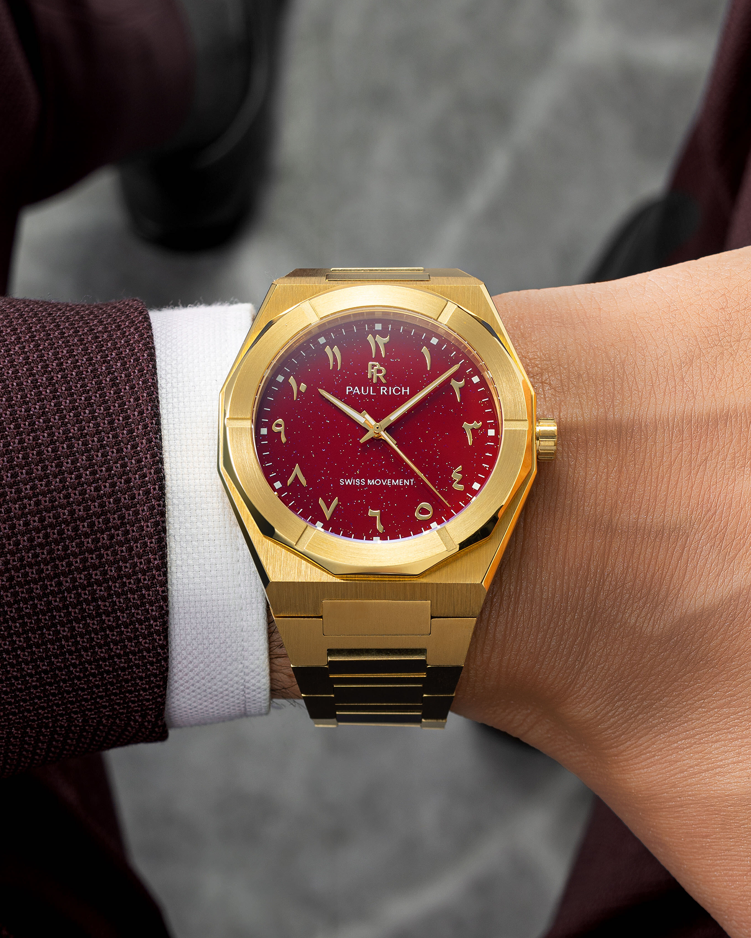 Paul Rich watch with brushed gold case and red aventurine dial featuring Arabic numerals, worn with a burgundy textured blazer.
