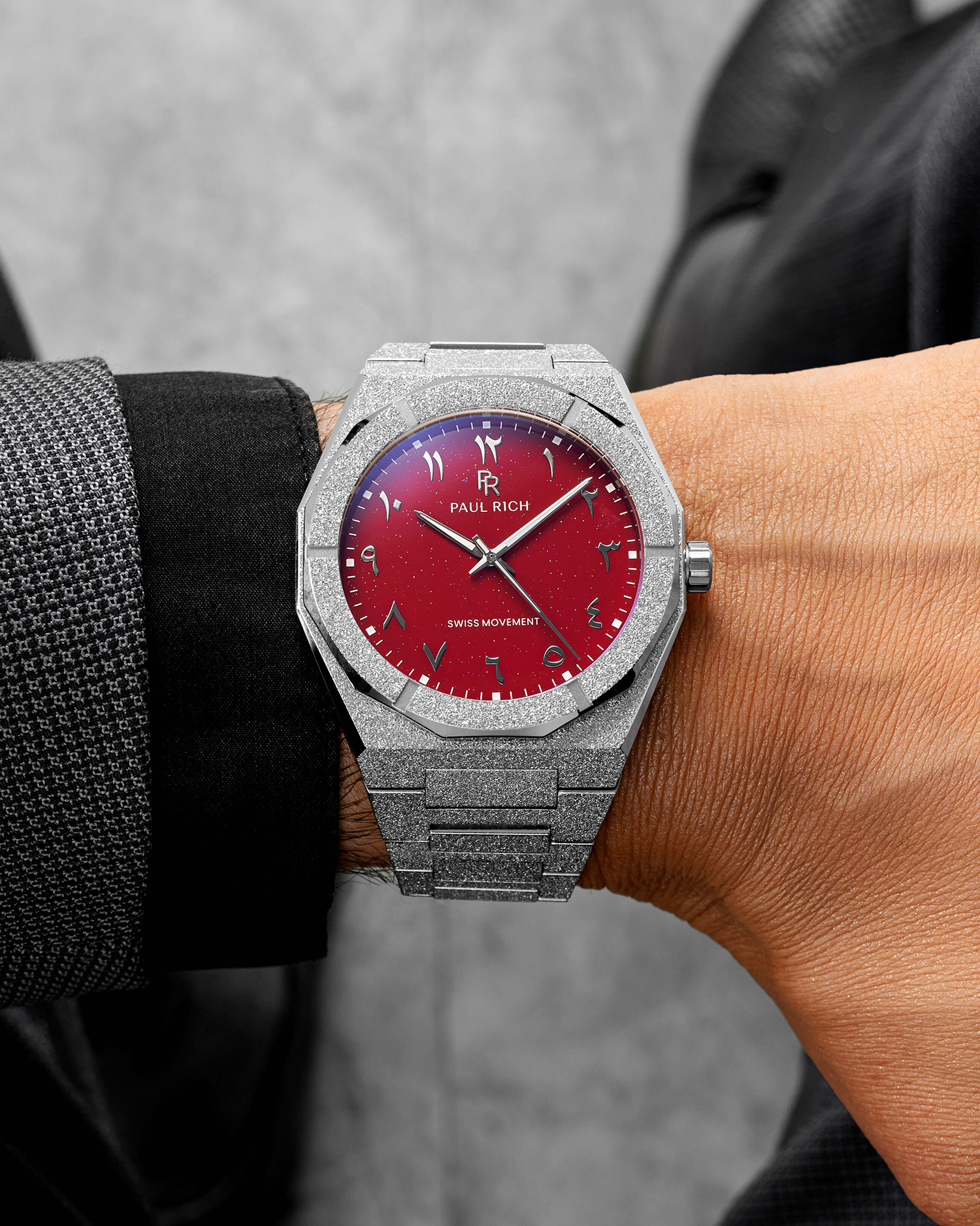 Paul Rich watch with frosted silver case and red aventurine dial with Arabic numerals, worn with a black and gray textured suit.