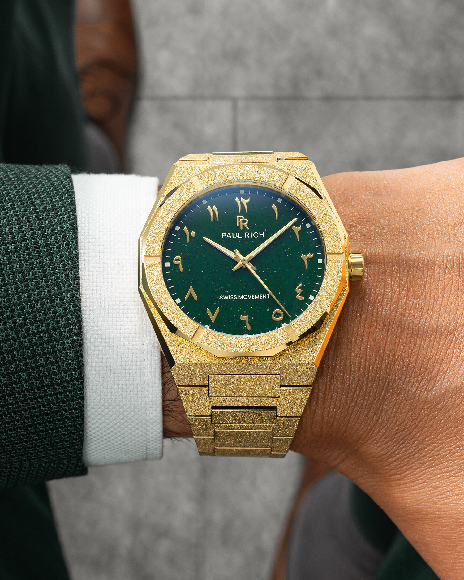 Paul Rich watch with frosted gold case and green aventurine dial with Arabic numerals, worn with a dark green textured suit and white cuff.