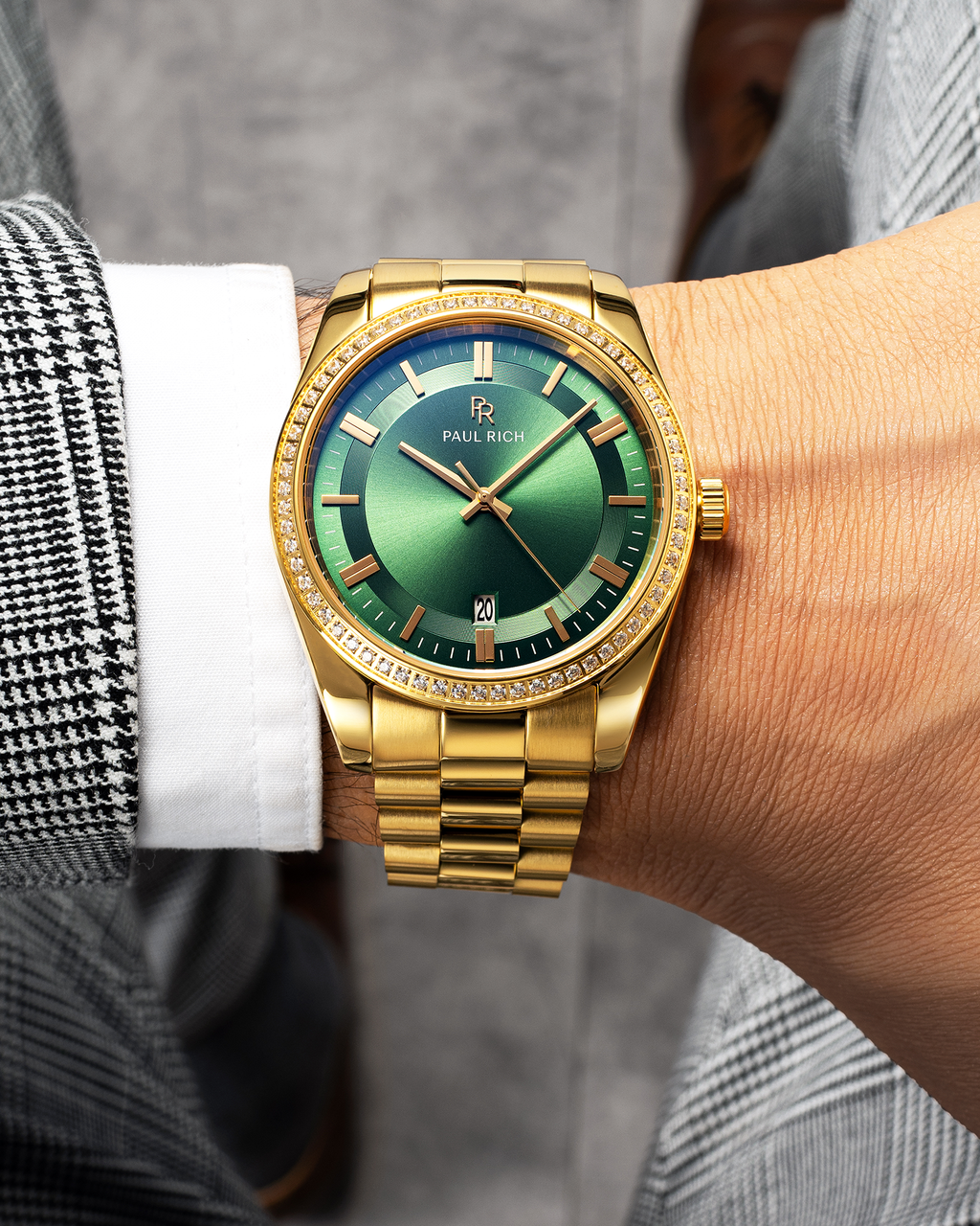 Gold-tone Paul Rich watch with green sunray dial, diamond-set bezel, and gold link bracelet, worn on wrist with a white shirt and black-and-white check blazer.