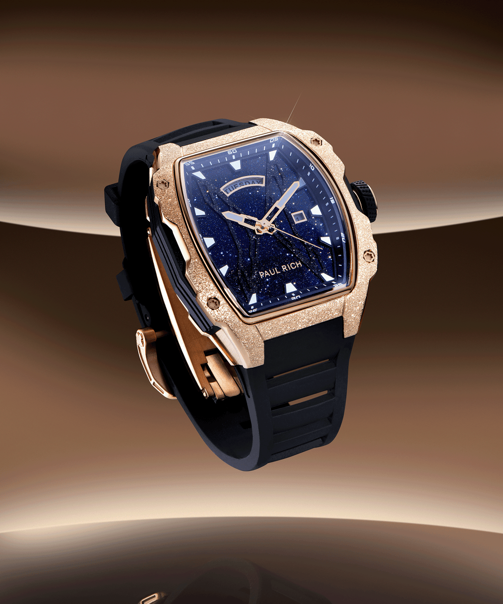 Paul Rich watch with frosted rose gold tonneau case, blue aventurine dial, day-date display, and black strap, floating over a bronze gradient background.