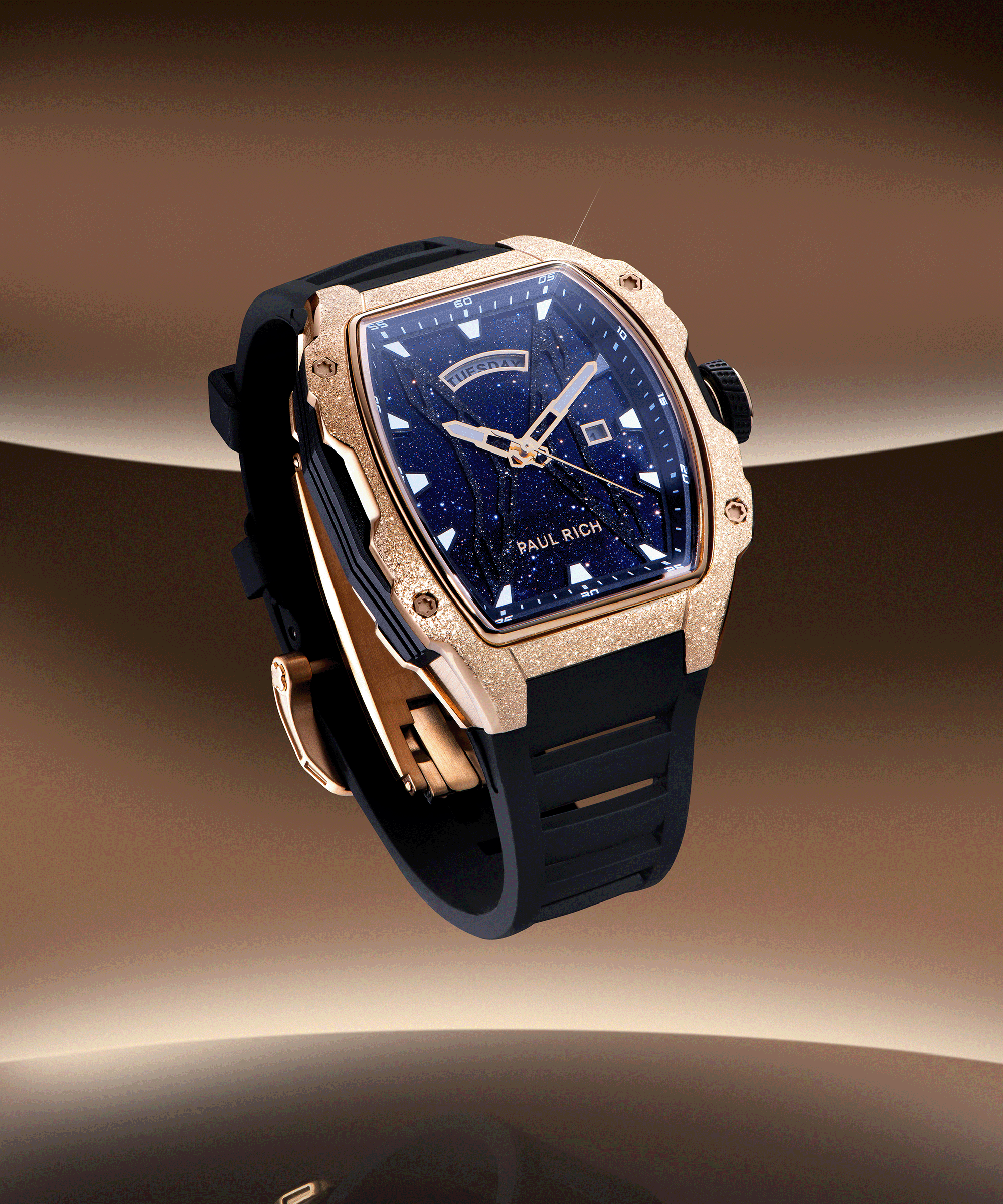 Paul Rich watch with frosted rose gold tonneau case, blue aventurine dial, day-date display, and black strap, floating over a bronze gradient background.