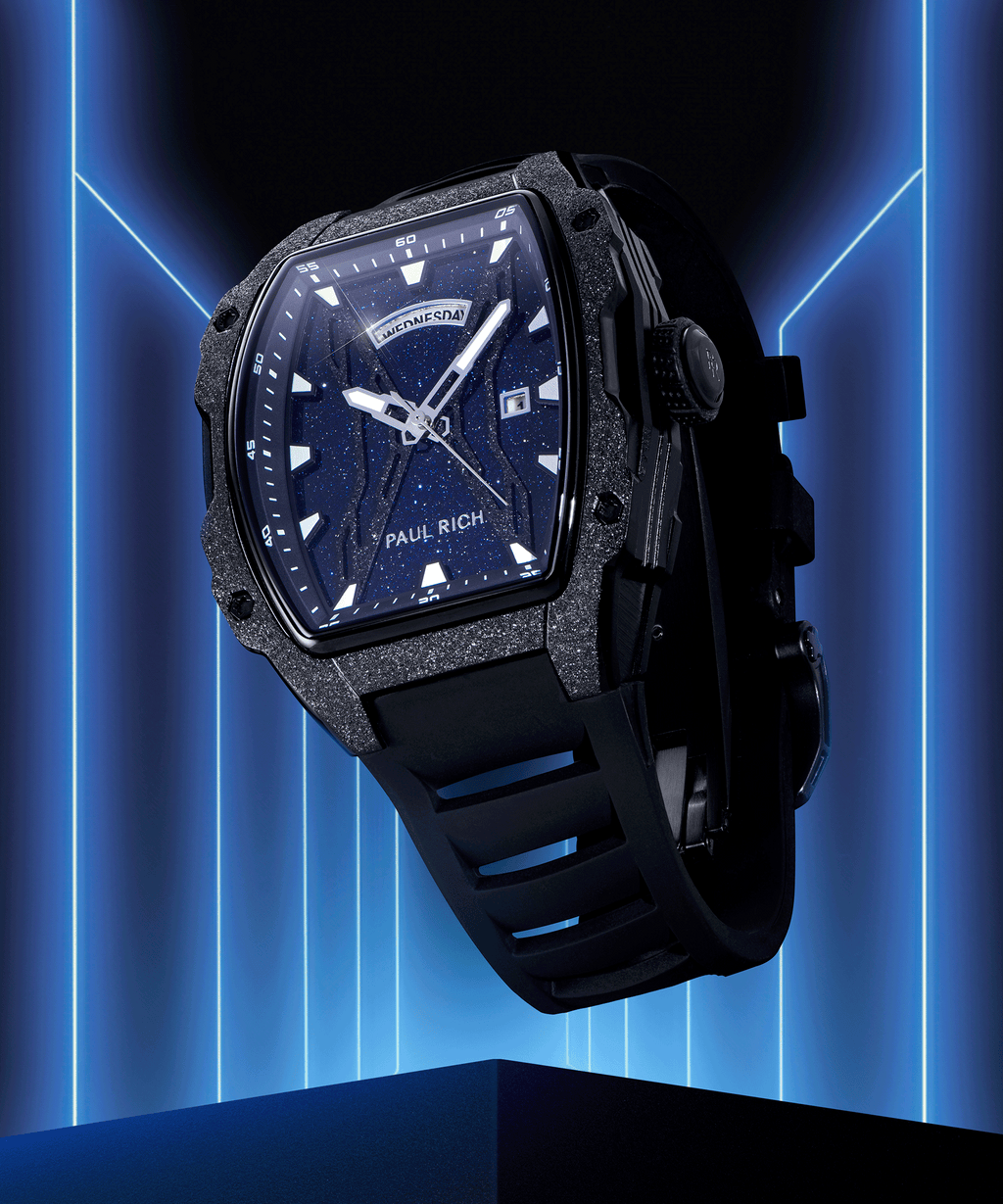 Paul Rich watch with blue aventurine dial, frosted bezel, day-date display, and black rubber strap, shown against neon blue backdrop.