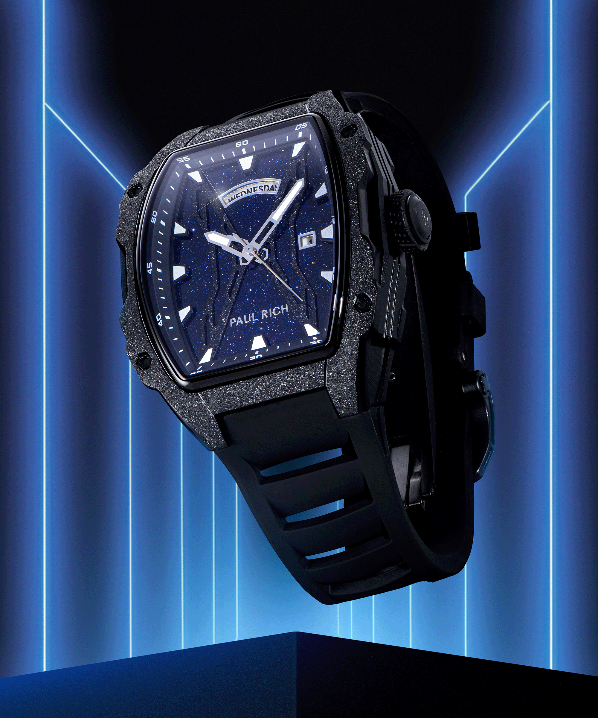 Paul Rich watch with blue aventurine dial, frosted bezel, day-date display, and black rubber strap, shown against neon blue backdrop.