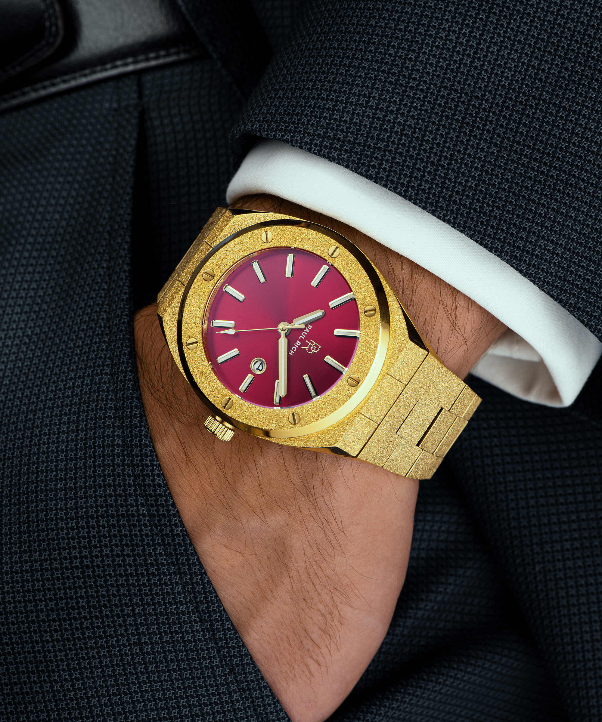 Paul Rich watch with frosted gold case and bracelet, metallic red sunray dial, and date window, worn on wrist with dark textured blazer and white cuff.