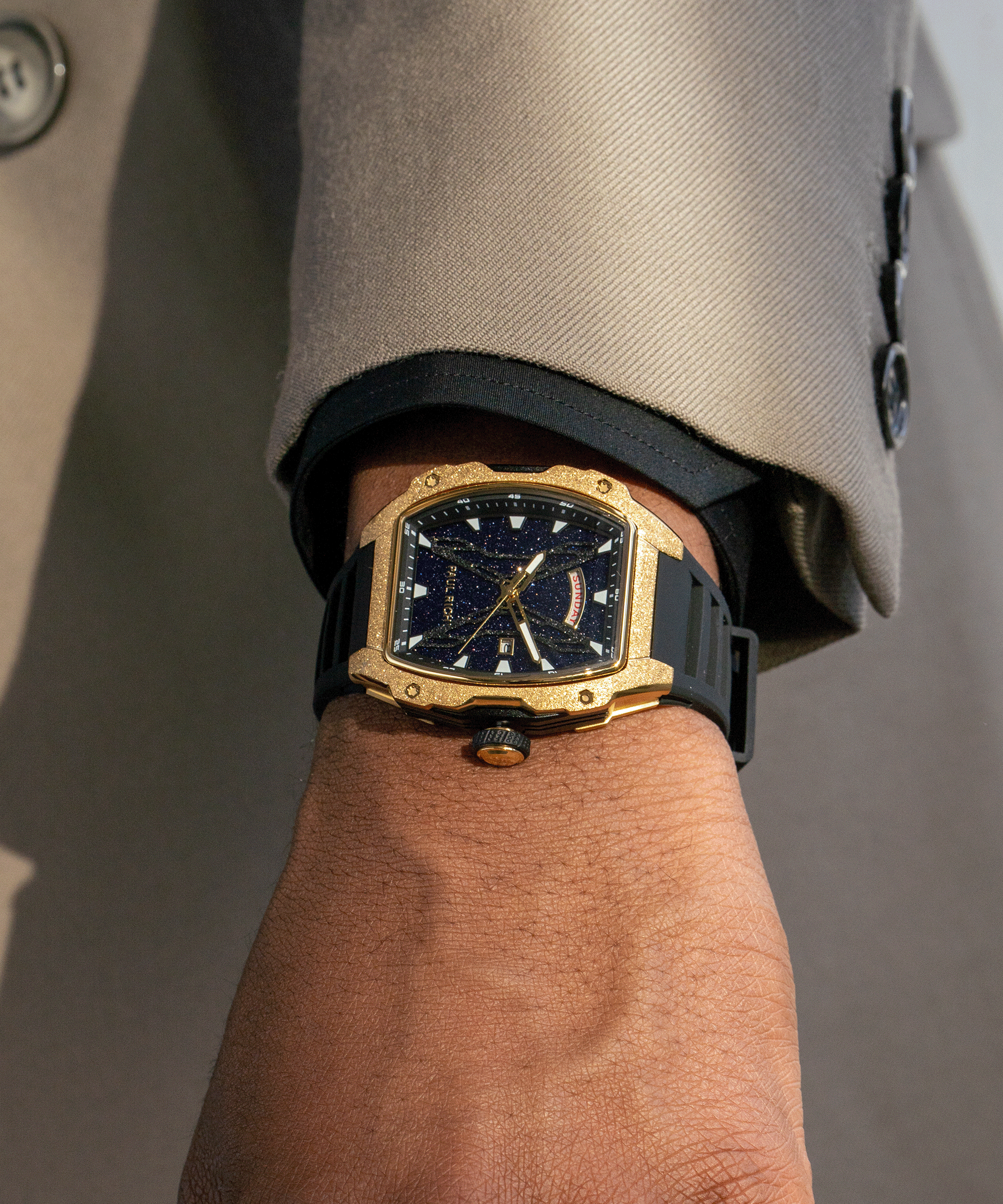 Man wearing a Paul Rich watch with frosted gold tonneau case, blue aventurine dial, day-date display, and black strap under beige suit sleeve.