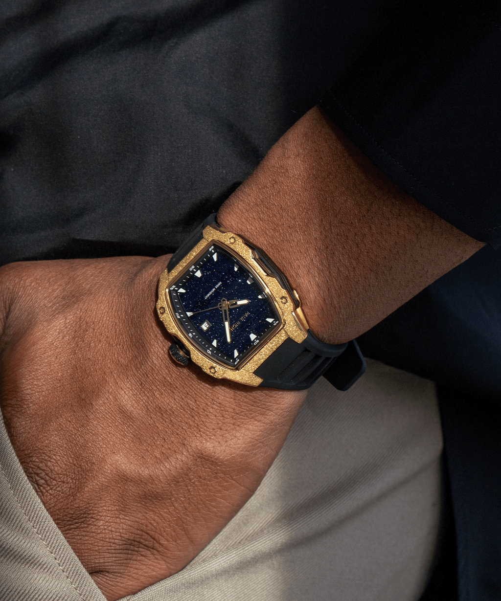 Tonneau-shaped Paul Rich watch with blue aventurine dial and gold frosted bezel worn on wrist, paired with beige pants and black top.