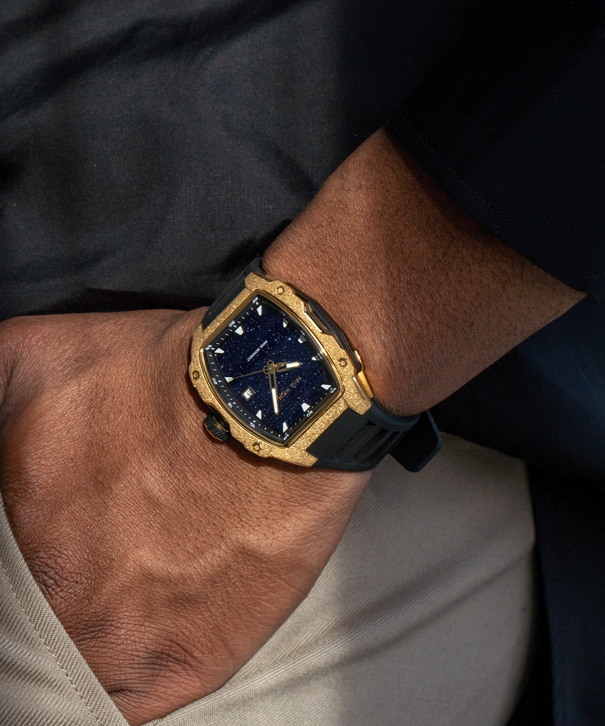 Tonneau-shaped Paul Rich watch with blue aventurine dial and gold frosted bezel worn on wrist, paired with beige pants and black top.