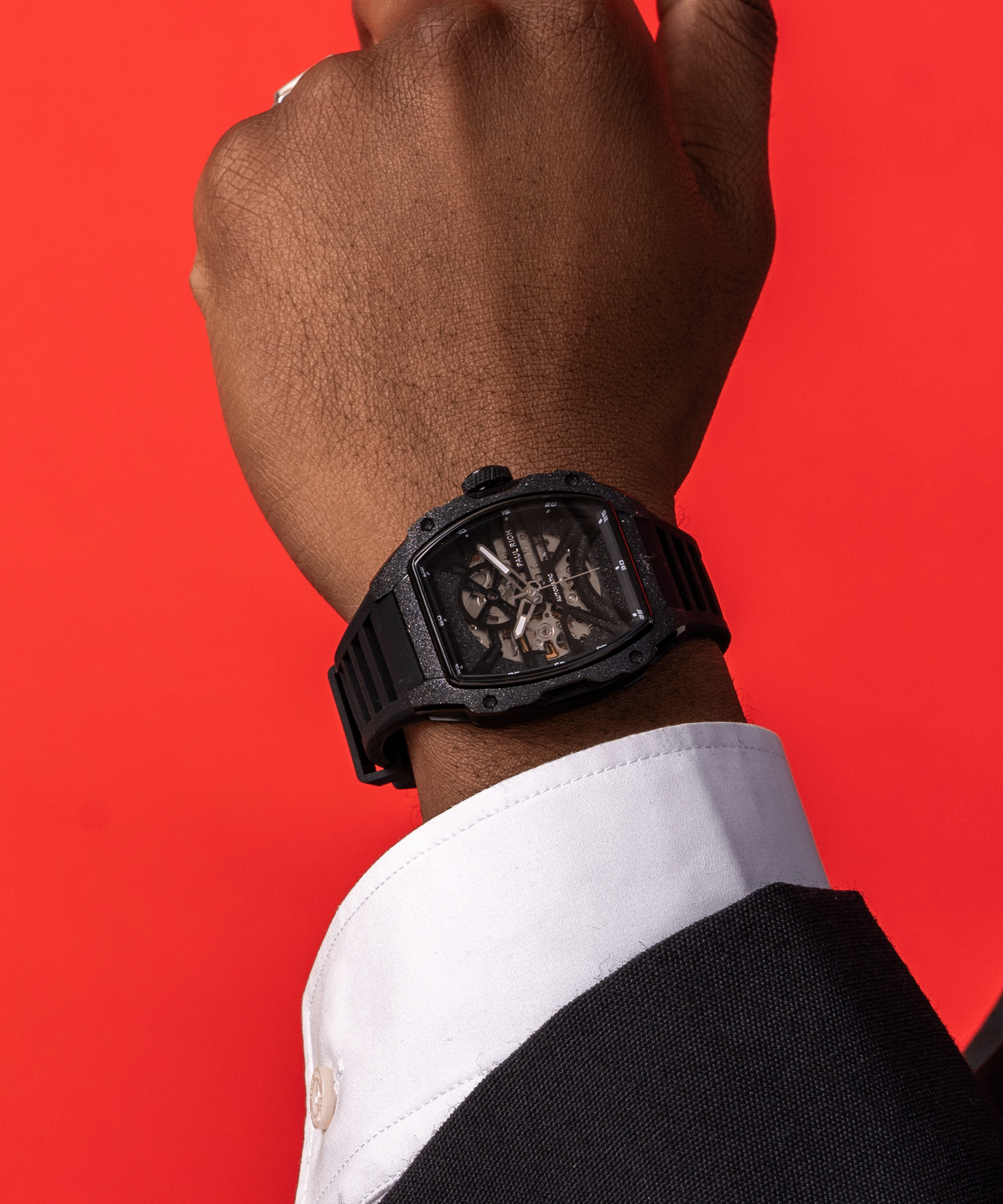 Man wearing a Paul Rich watch with frosted black tonneau case and skeleton dial, paired with black suit and white shirt, against red background.