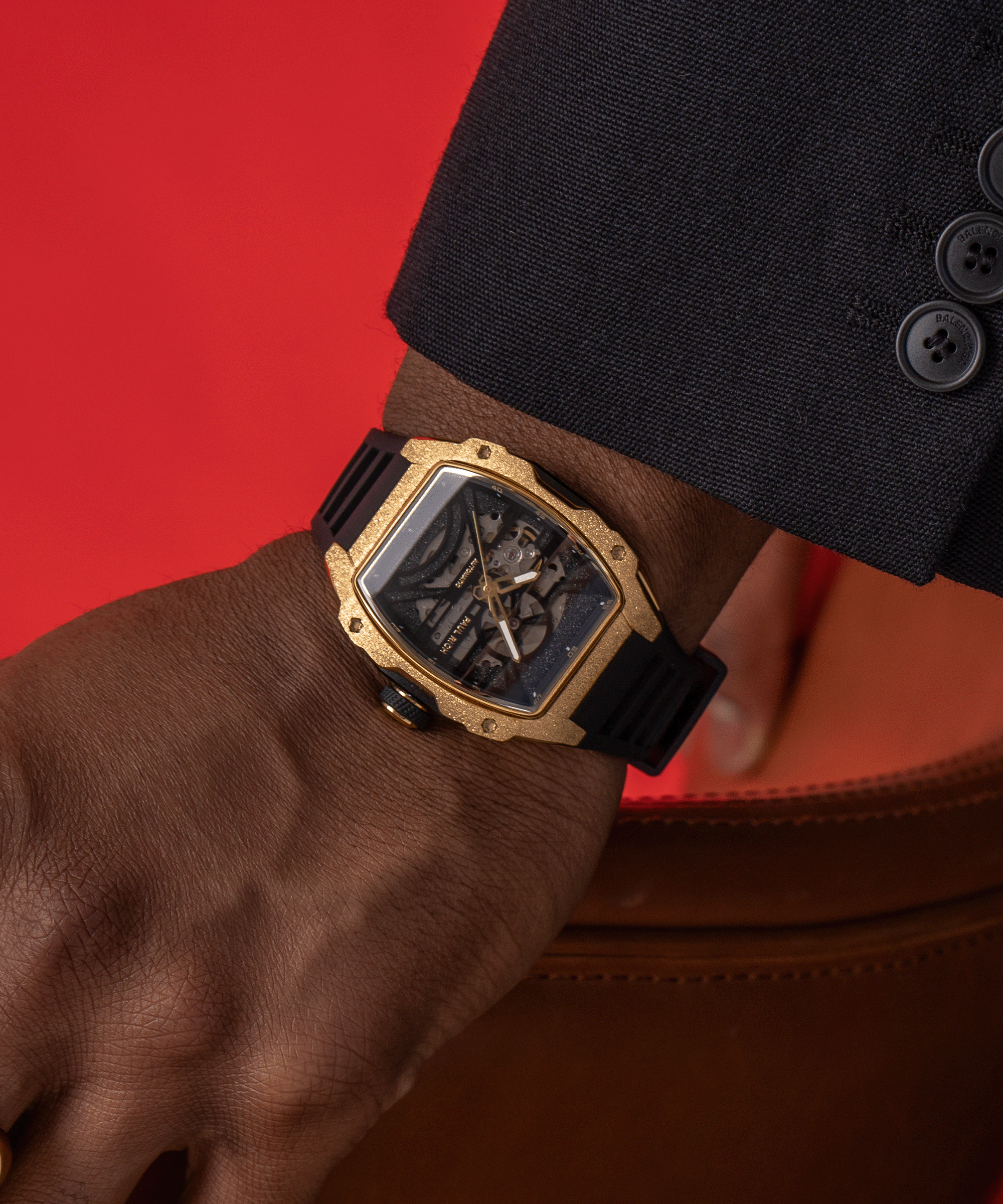 Man wearing a Paul Rich watch with frosted gold tonneau case and black skeleton dial, paired with black suit, against a red backdrop.