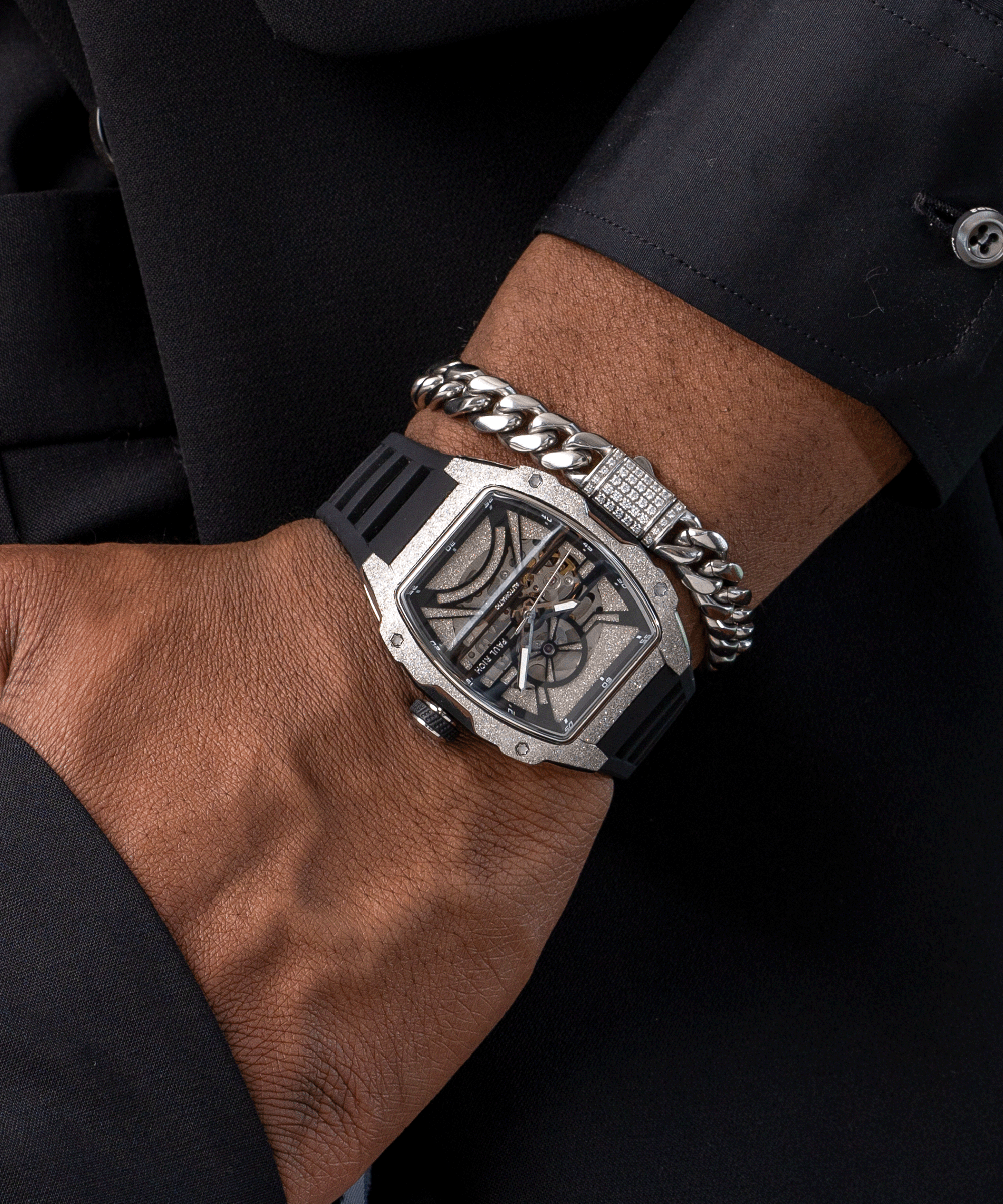Man wearing a Paul Rich watch with frosted silver tonneau case and skeleton dial, paired with silver Cuban link bracelet over black suit.