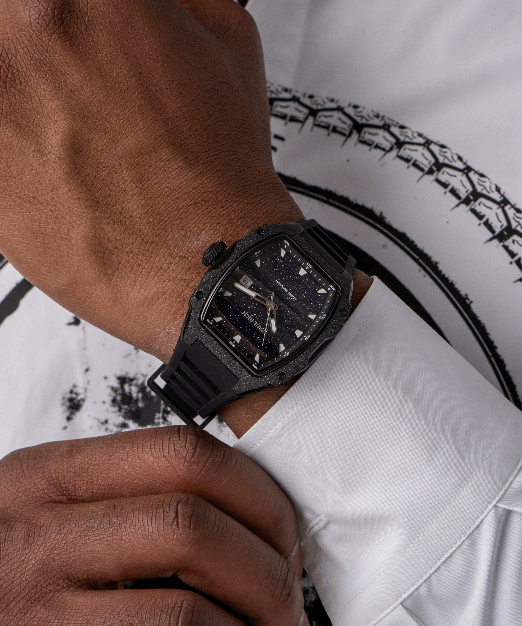 Tonneau-shaped Paul Rich watch with blue aventurine dial and black frosted bezel worn on wrist with white shirt and graphic print.