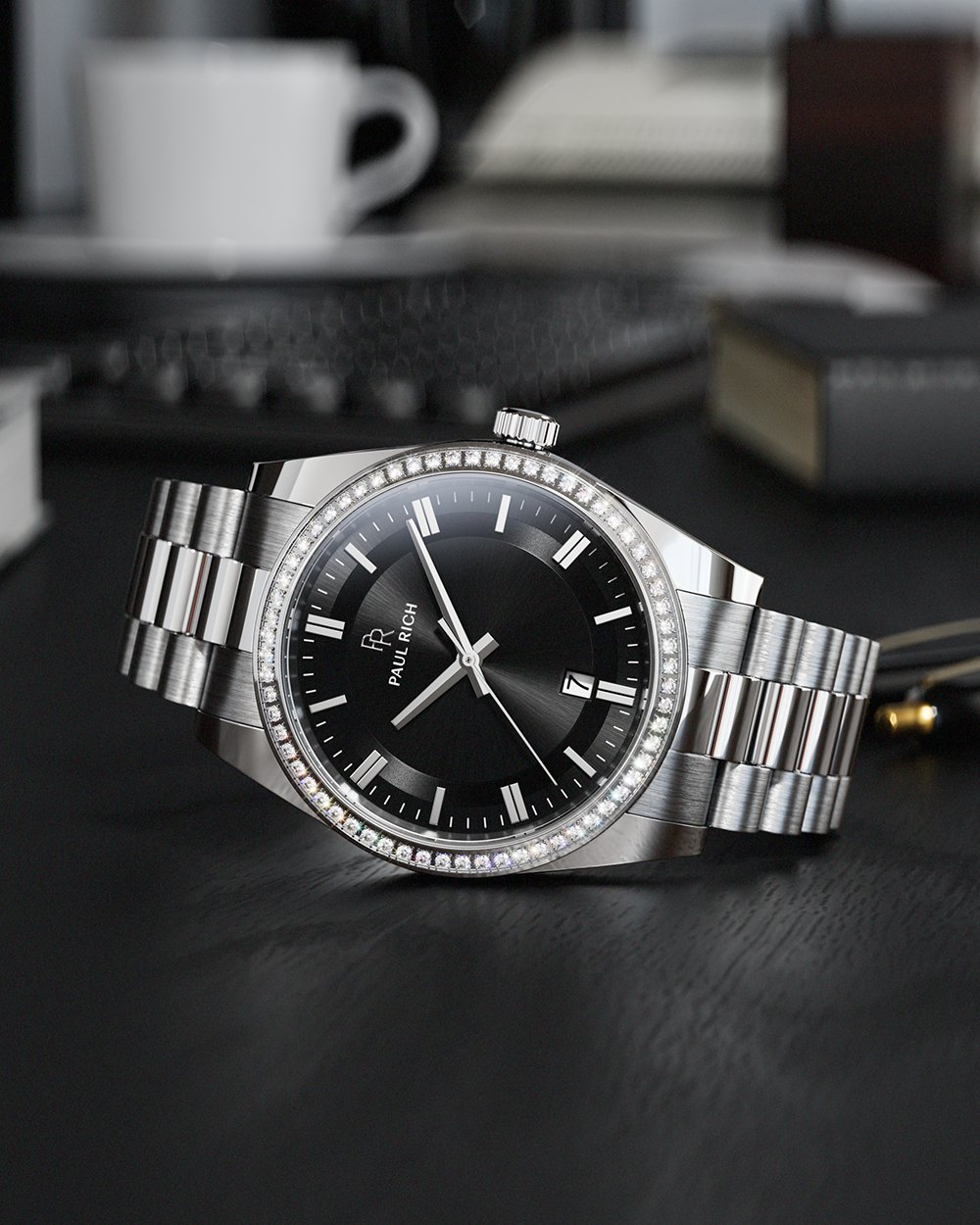 Silver-tone Paul Rich watch with black sunray dial, diamond-set bezel, and silver link bracelet, displayed on a black desk surface with a white coffee cup, keyboard, and books blurred in the background.