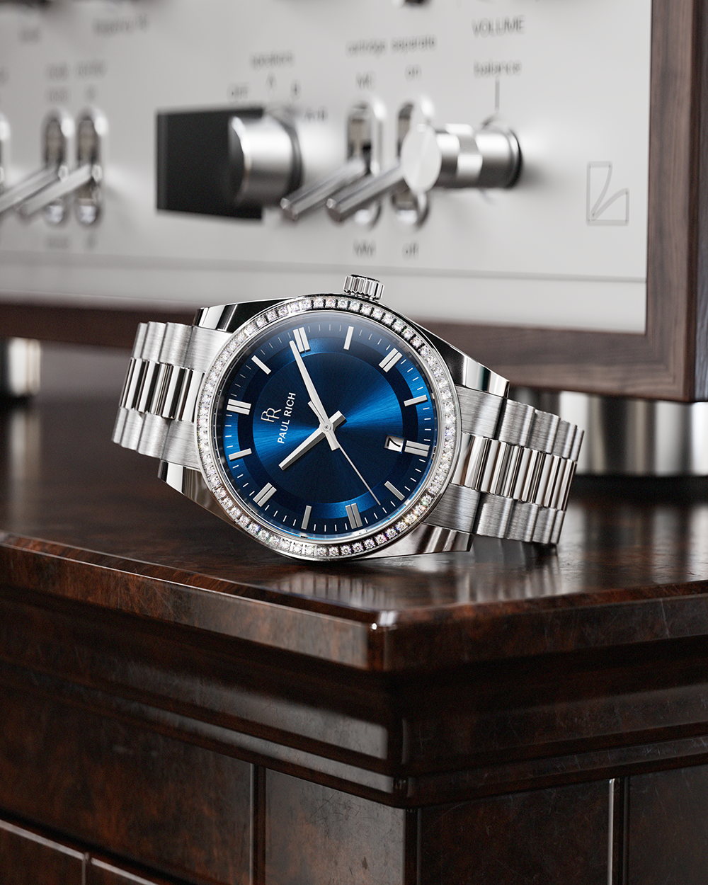 Silver-tone Paul Rich watch with blue sunray dial, diamond-set bezel, and silver link bracelet, displayed on a dark wood cabinet with a vintage silver audio receiver in the background.