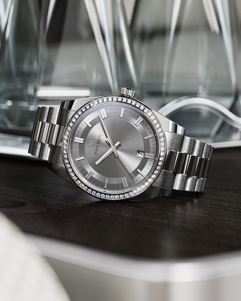 Silver-tone Paul Rich watch with silver sunray dial, diamond-set bezel, and silver link bracelet, displayed on a dark wooden surface with reflective glass decanters in the background.