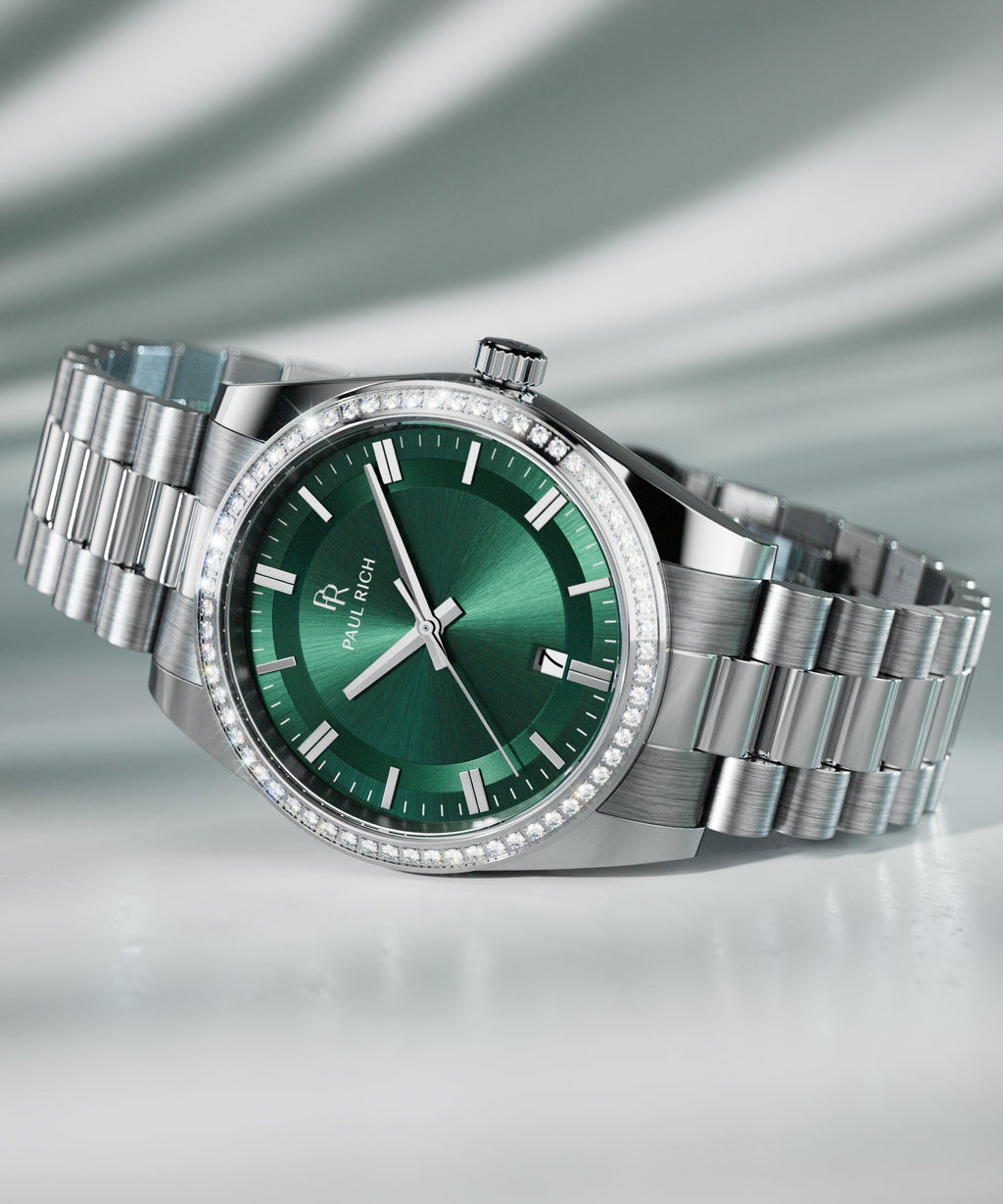 Silver wristwatch with a green sunburst dial, crystal-set bezel, and metal bracelet displayed on a reflective surface with soft lighting.