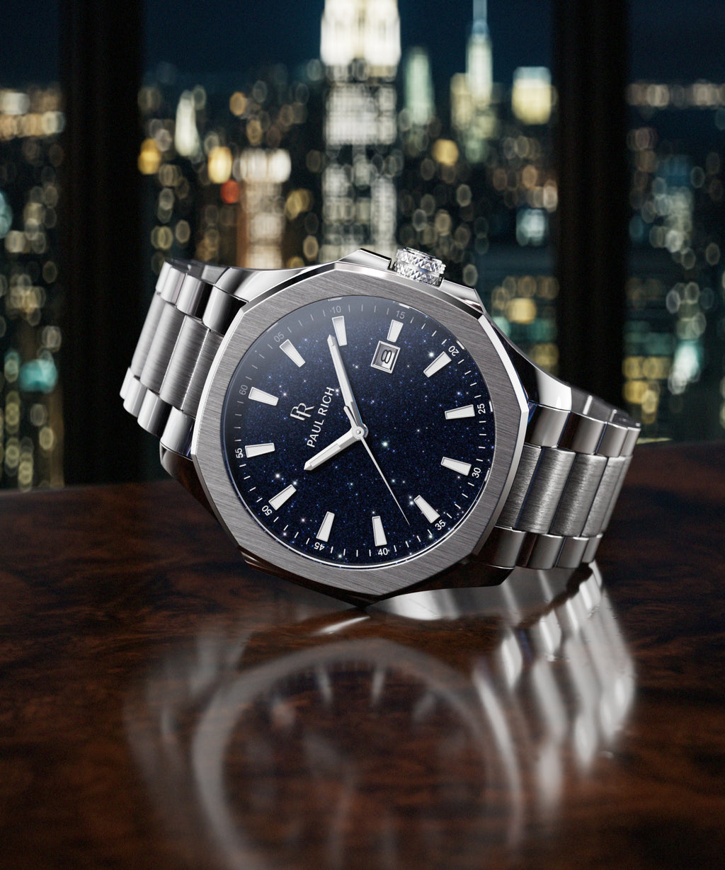 Paul Rich watch with brushed silver case and blue aventurine dial resting on dark wood surface, with NYC skyline at night in background.