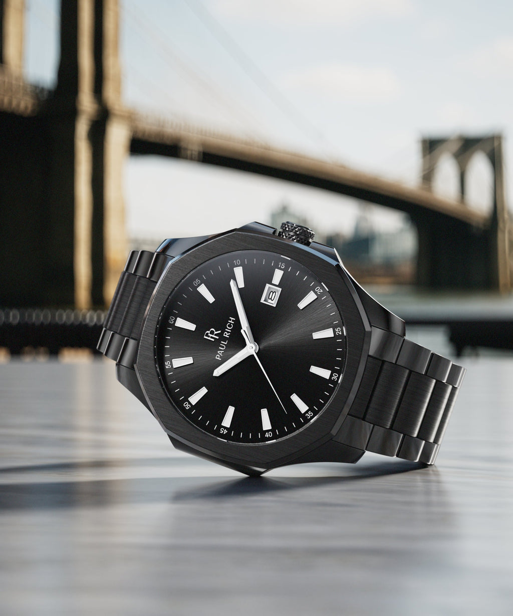 Black Paul Rich watch with a sunray dial and steel bracelet, styled on a table with the Brooklyn Bridge in the background.