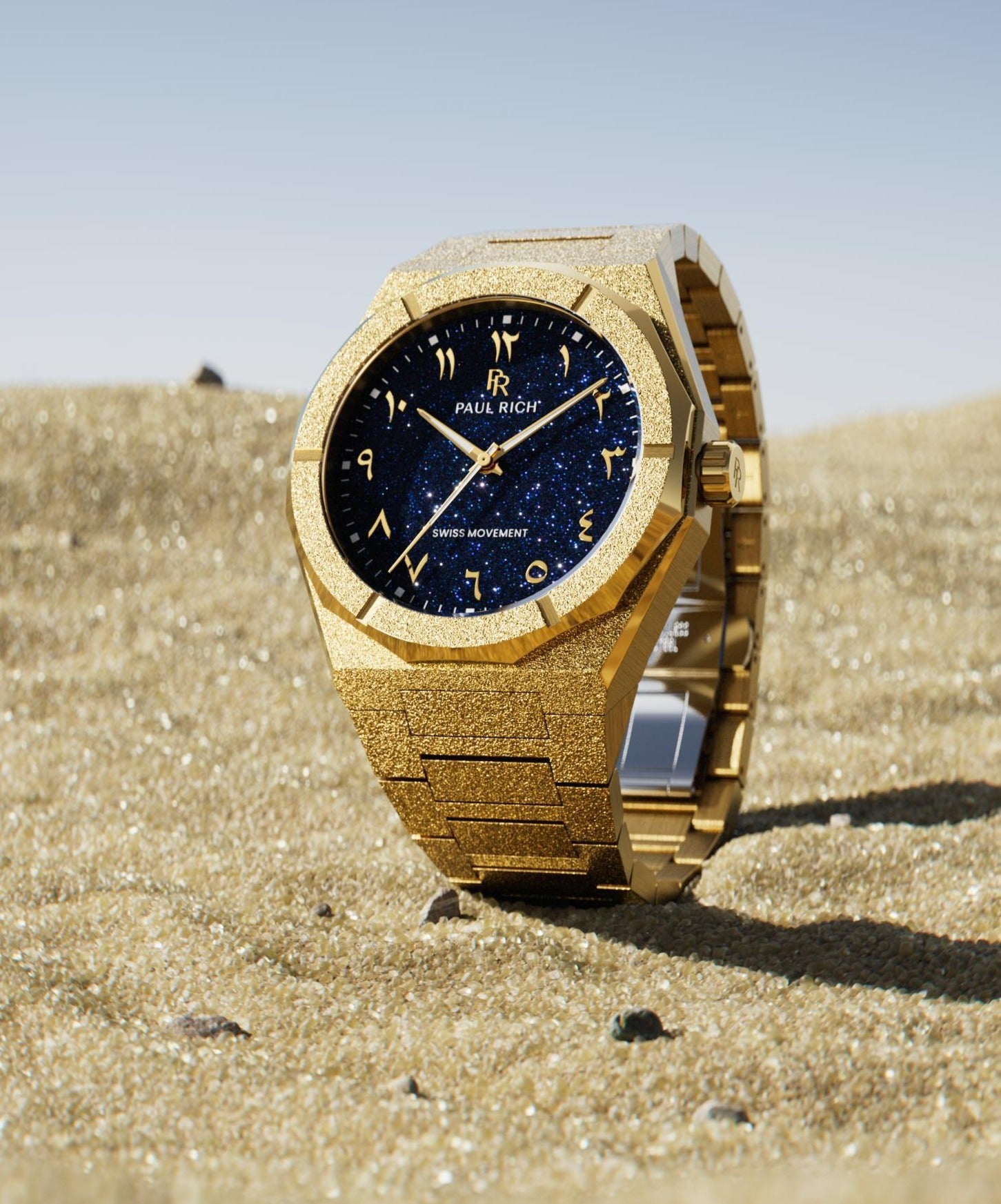 Frosted gold watch with a blue aventurine dial and Arabic numerals, standing upright on sunlit sand with a clear blue sky in the background.