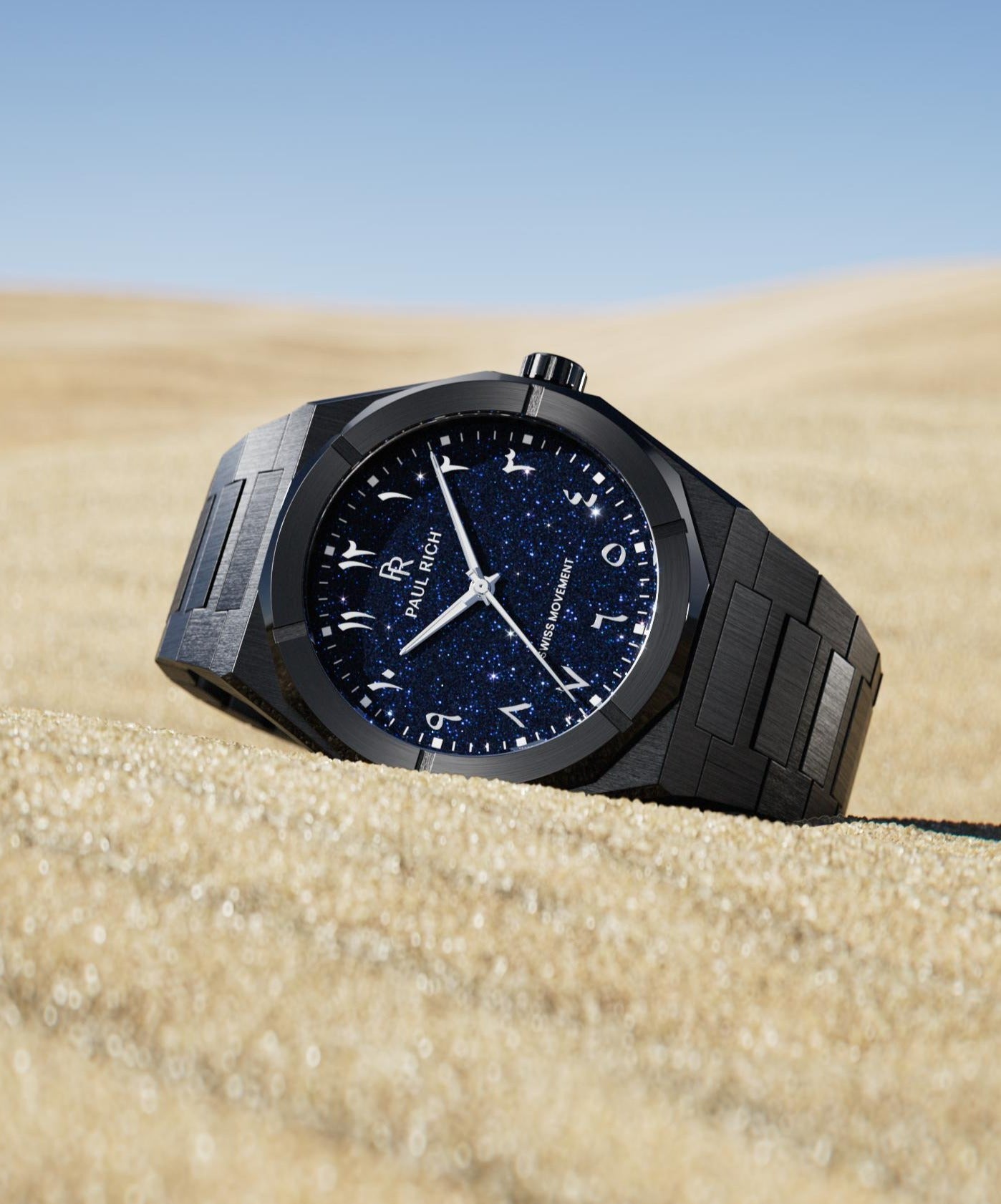 Paul Rich watch with brushed black case and blue aventurine dial with Arabic numerals, displayed on golden sand under a clear sky.