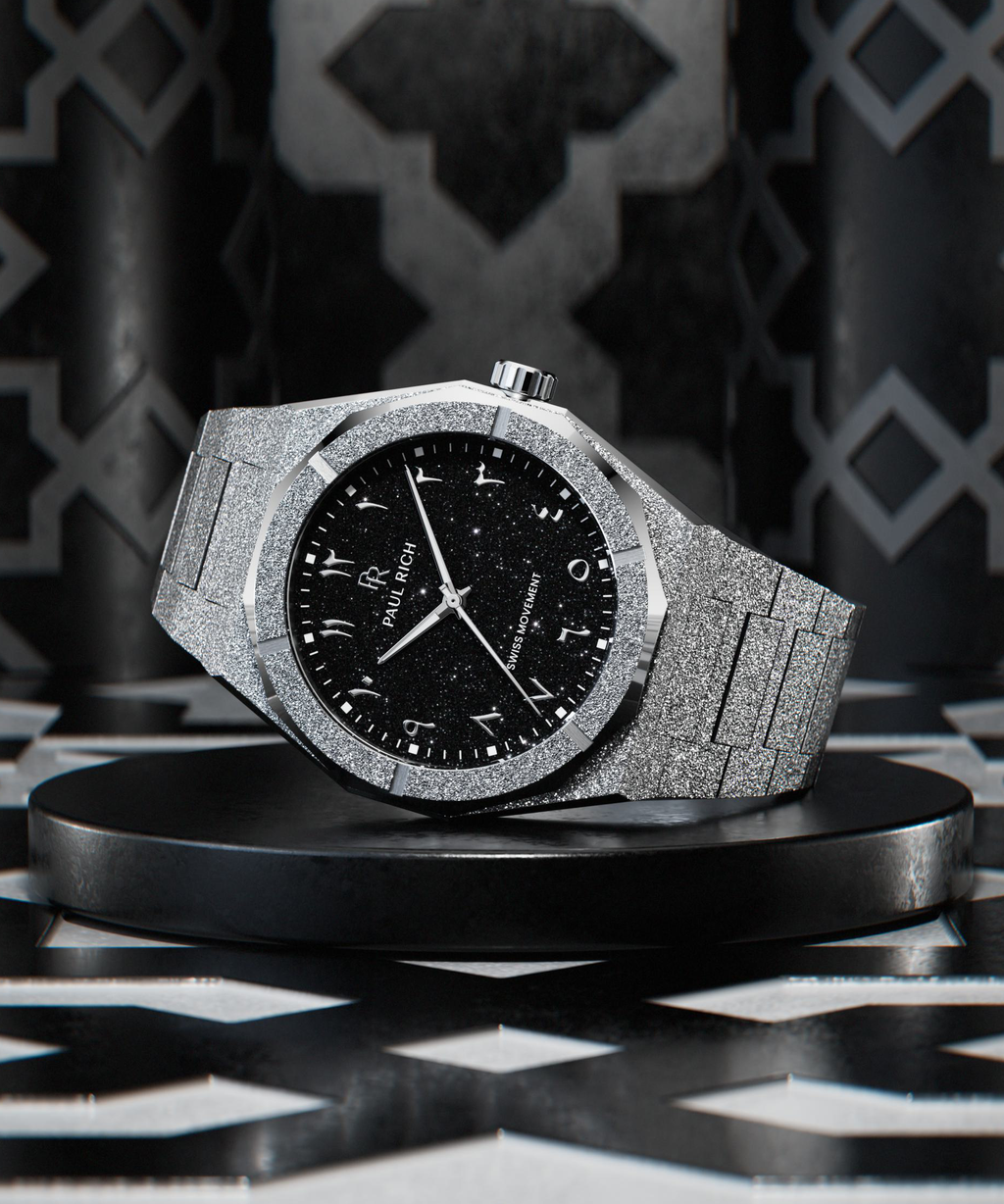 Silver wristwatch with a frosted case and bracelet, black aventurine dial, and silver Arabic numerals displayed on a black platform with geometric patterned background.