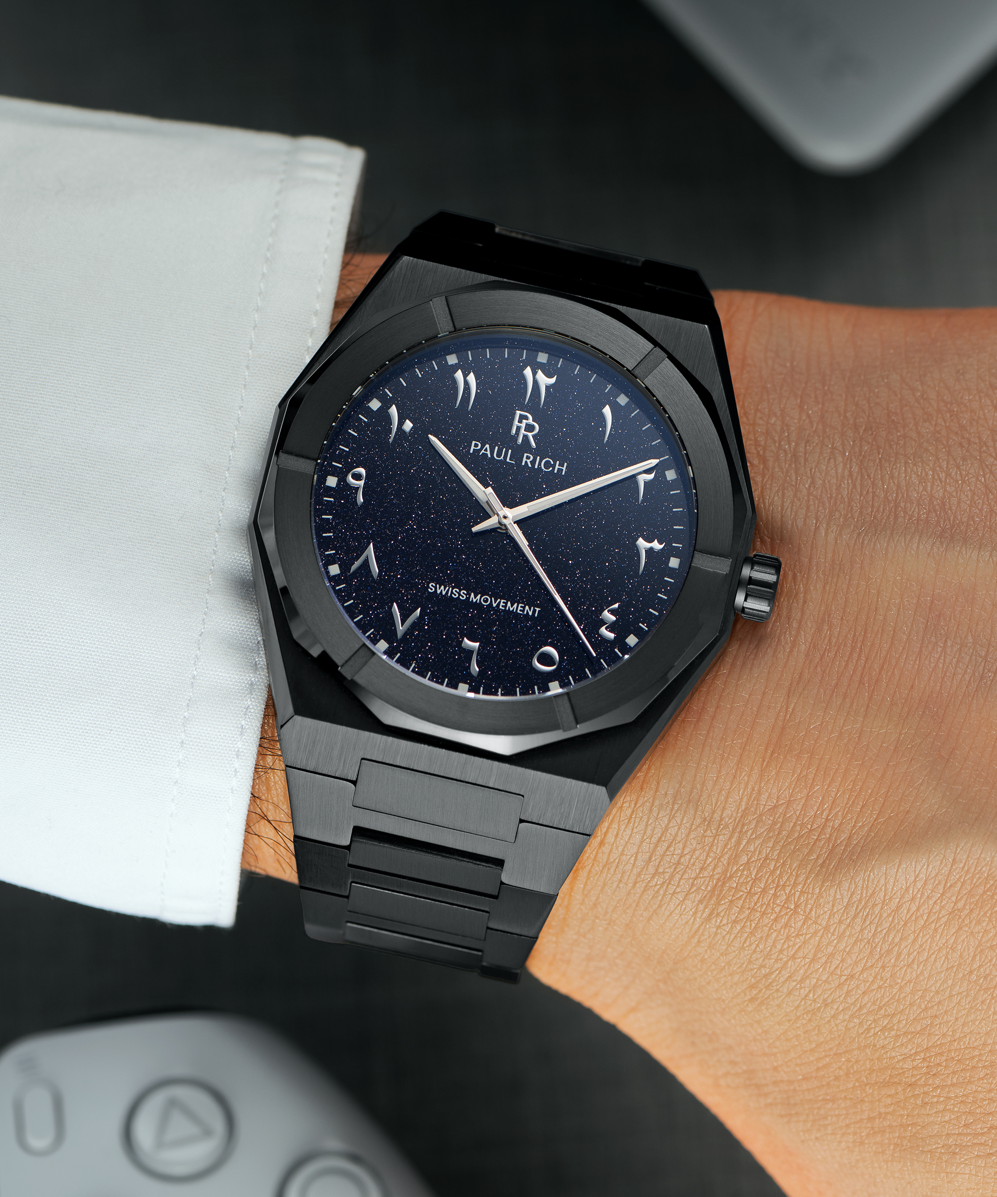 Paul Rich watch with brushed black case and blue aventurine dial with Arabic numerals, worn with a white cuff over a dark surface.