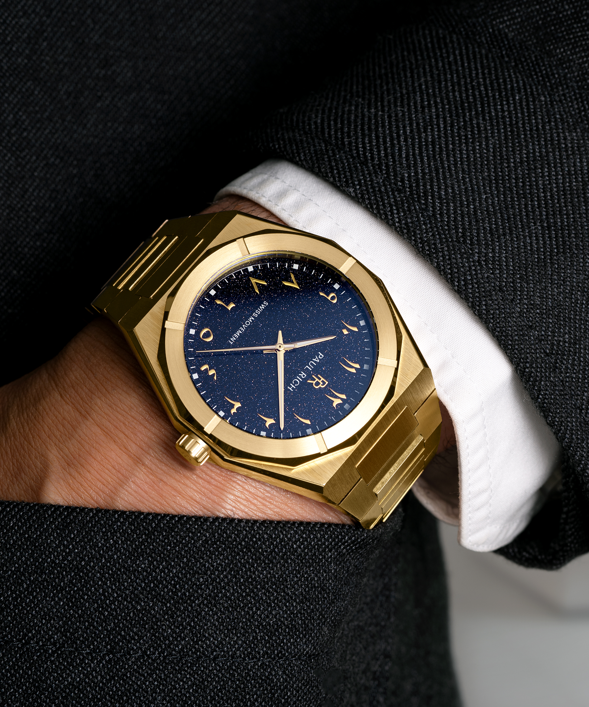 Paul Rich watch with brushed gold case and blue aventurine dial with Arabic numerals, worn with a black suit and white cuff, hand in pocket.