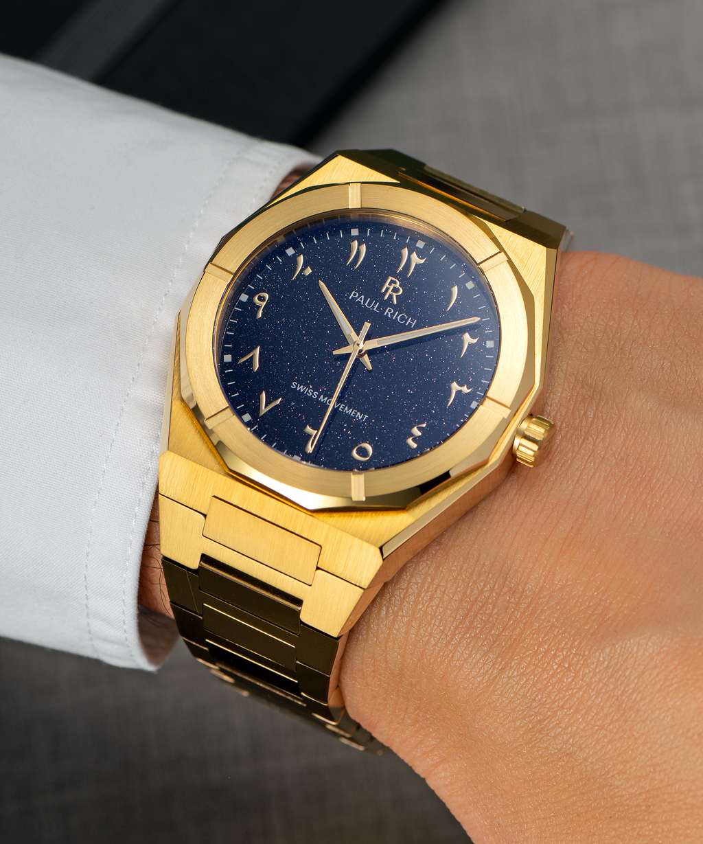 Paul Rich watch with brushed gold case and blue aventurine dial with Arabic numerals, worn with a white cuff over a grey suit.