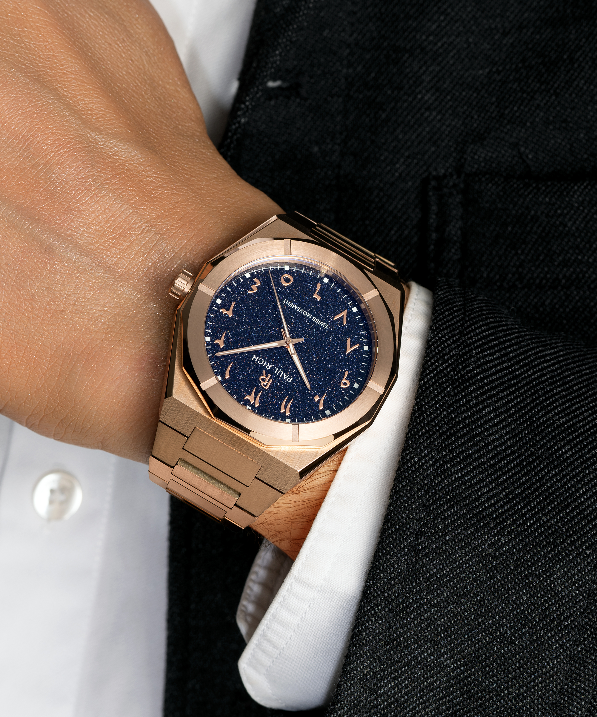 Paul Rich watch with brushed rose gold case and blue aventurine dial with Arabic numerals, worn with a white cuff and black suit.