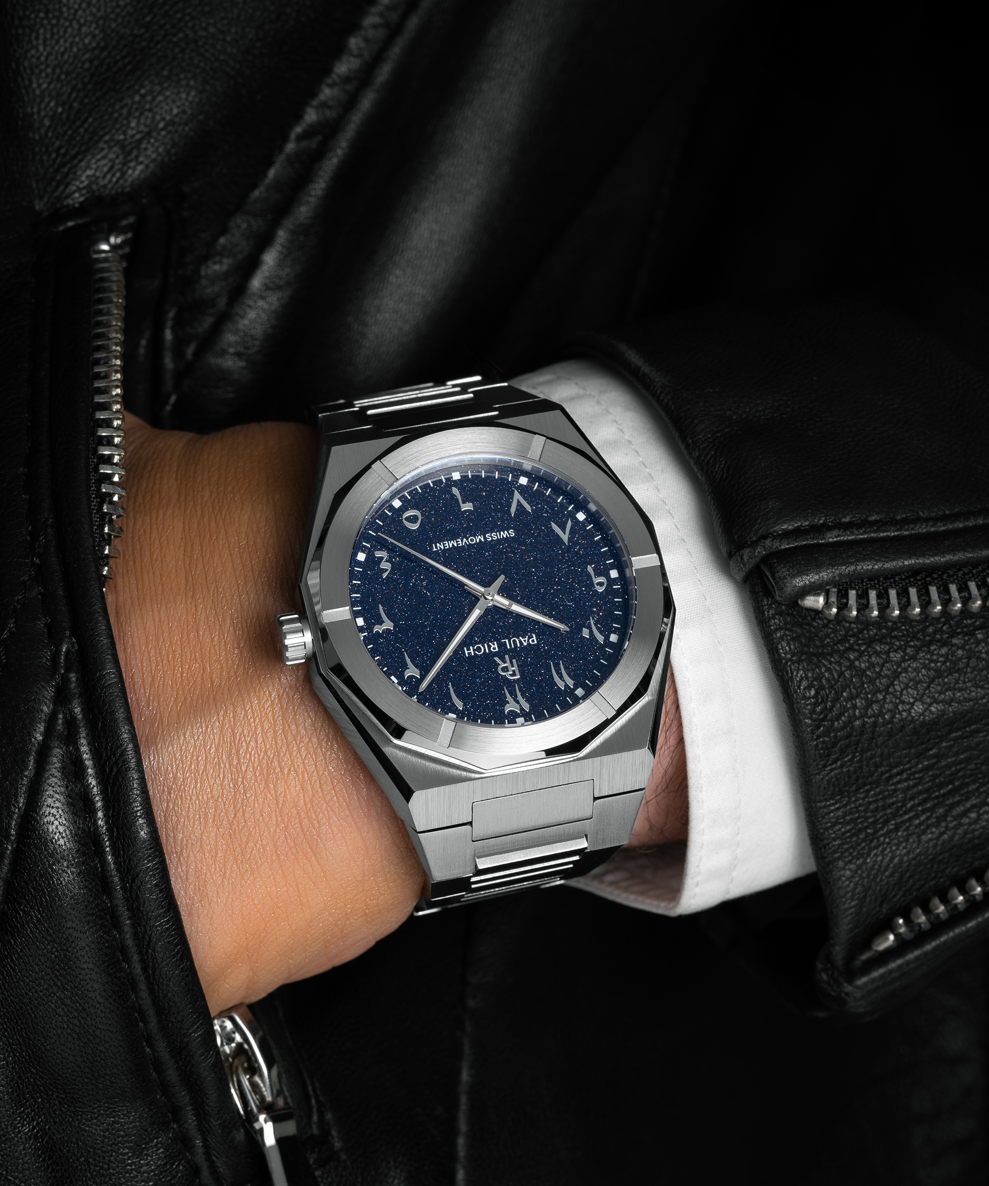 Paul Rich watch with brushed silver case and blue aventurine dial with Arabic numerals, worn with a white cuff and black leather jacket.