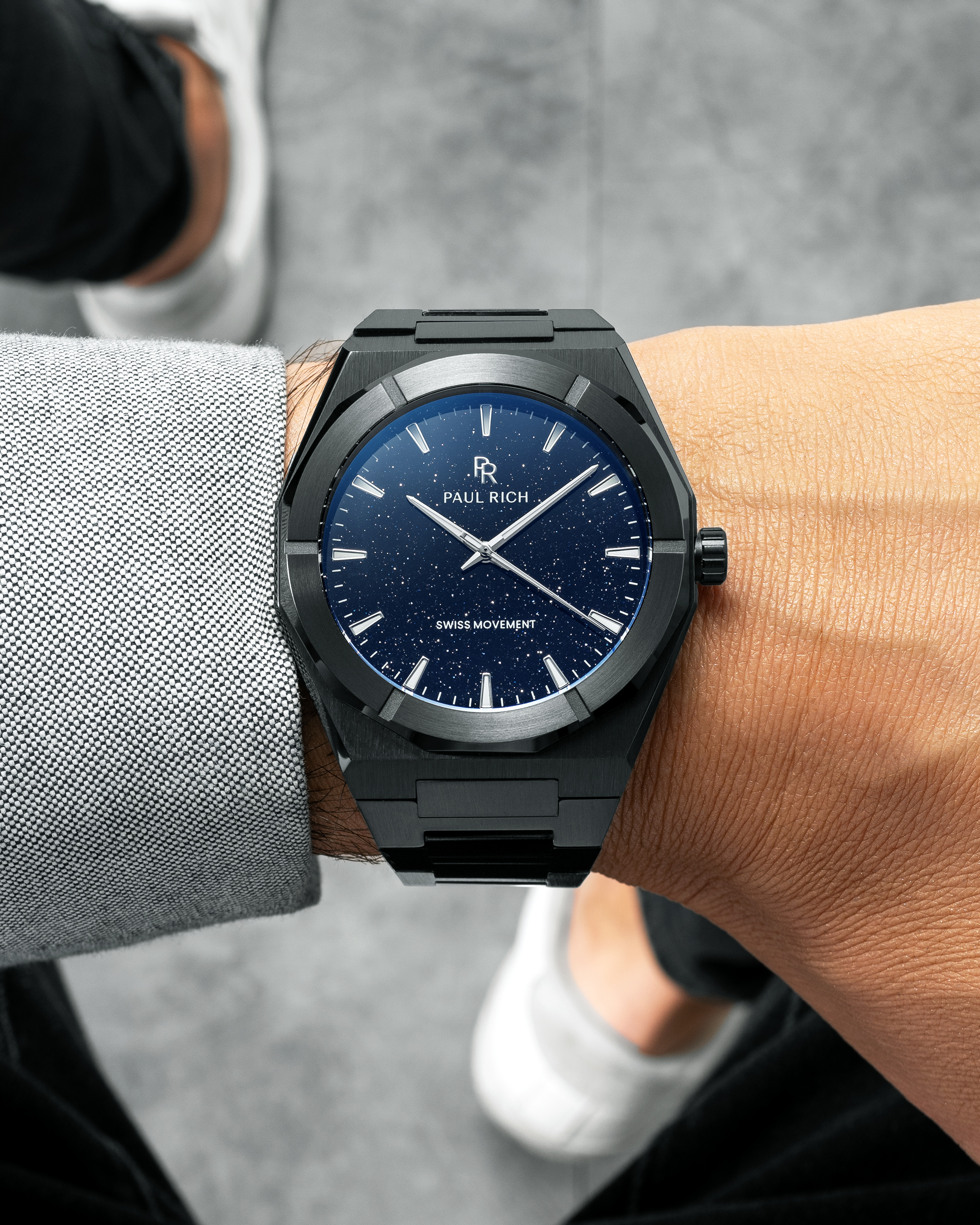 Black Paul Rich watch with blue aventurine dial worn on wrist, paired with light grey shirt and black pants, white sneakers visible.
