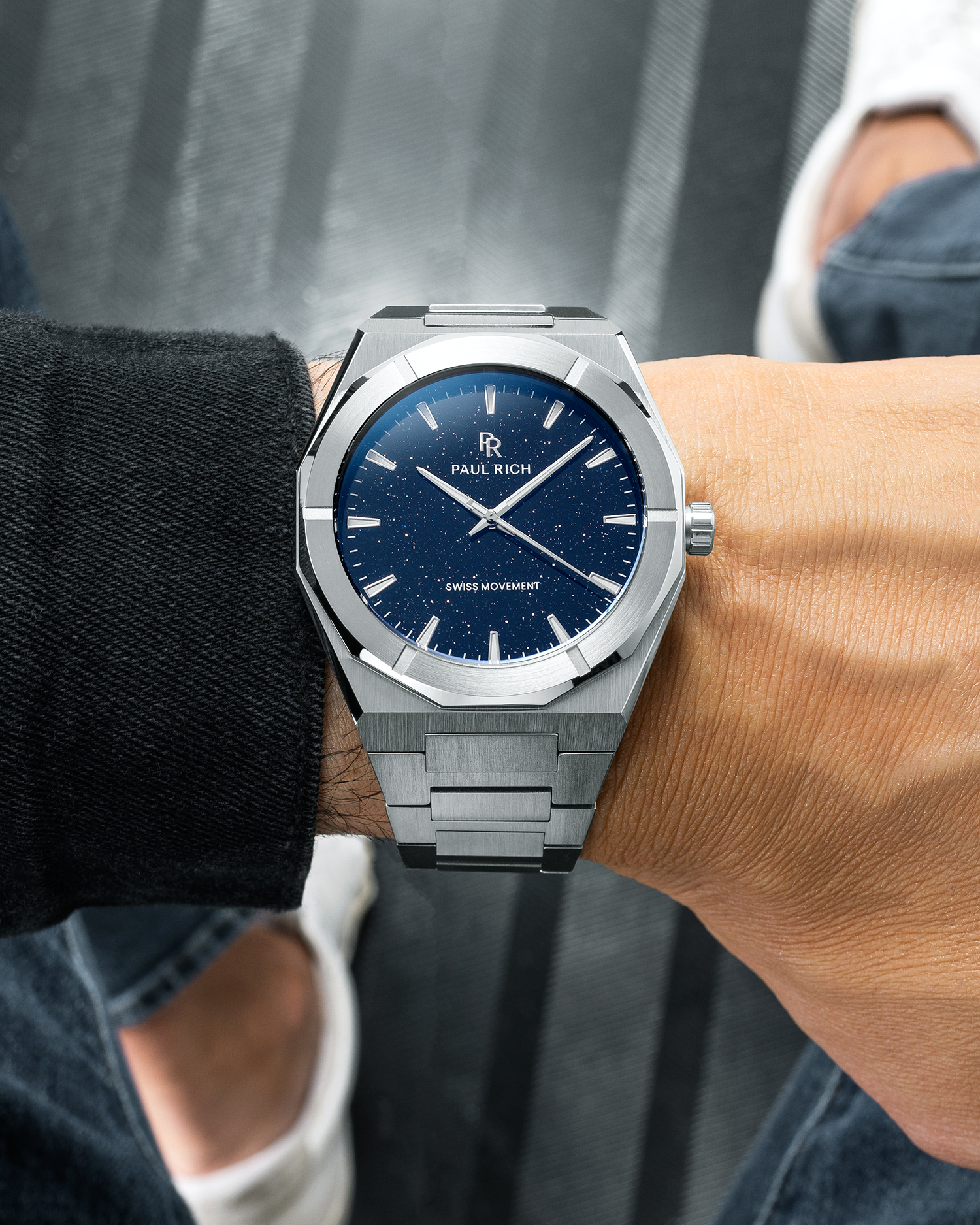 Silver Paul Rich watch with blue aventurine dial on wrist, worn over black sleeve with jeans and white sneakers visible.