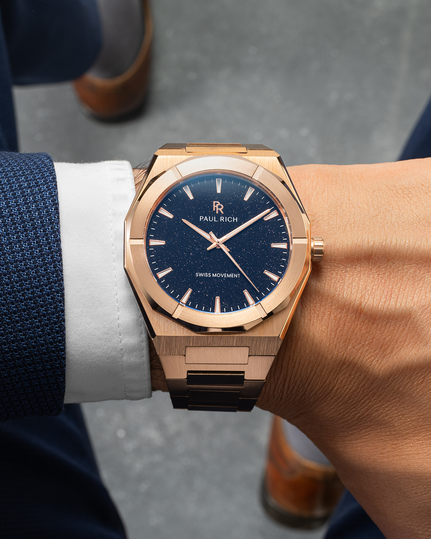Rose gold Paul Rich watch with blue aventurine dial worn on wrist, paired with navy suit and white shirt, brown shoes in background.