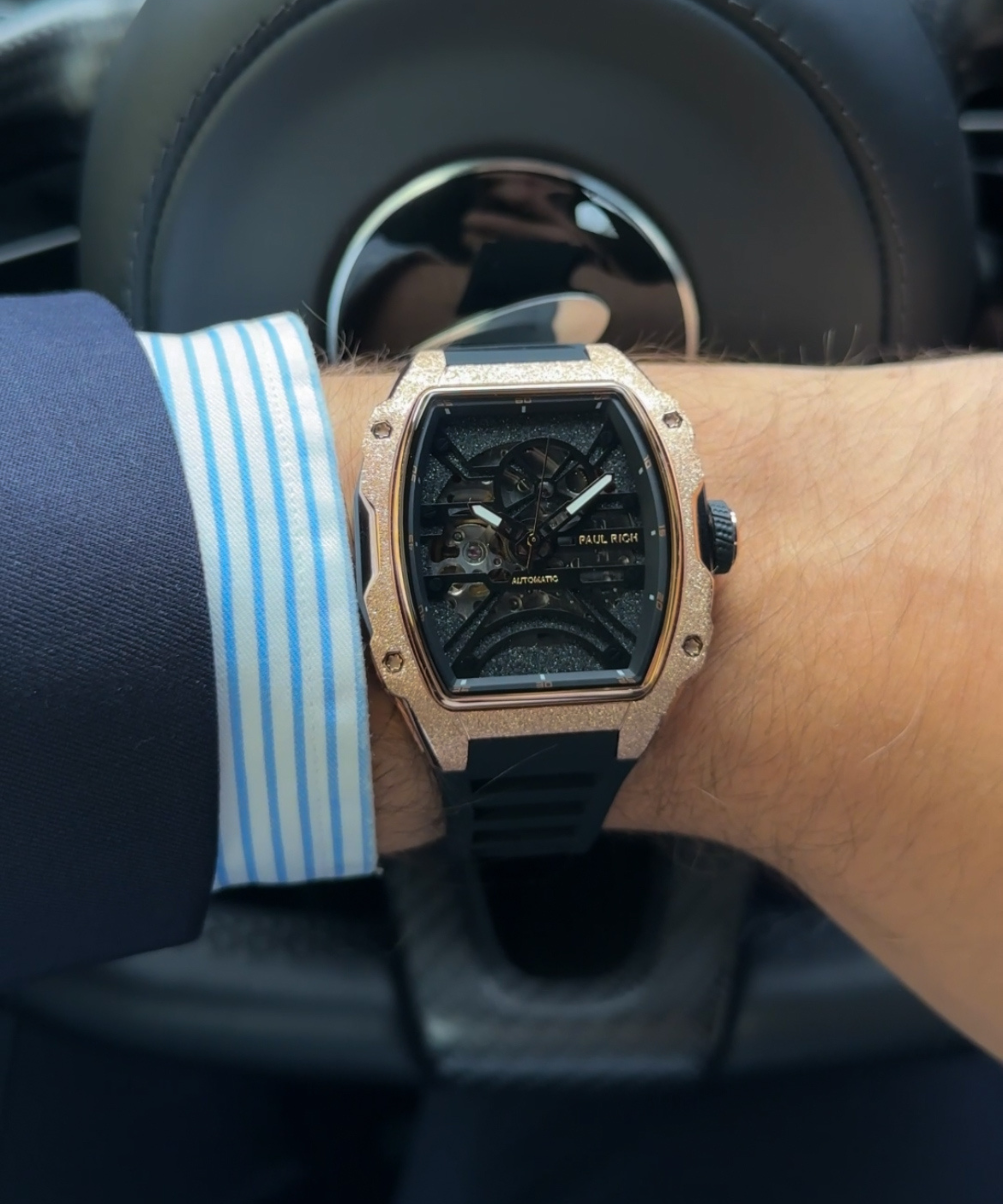 Man wearing a Paul Rich watch with frosted rose gold case and black skeleton dial, seen over a cuffed dress shirt while driving.