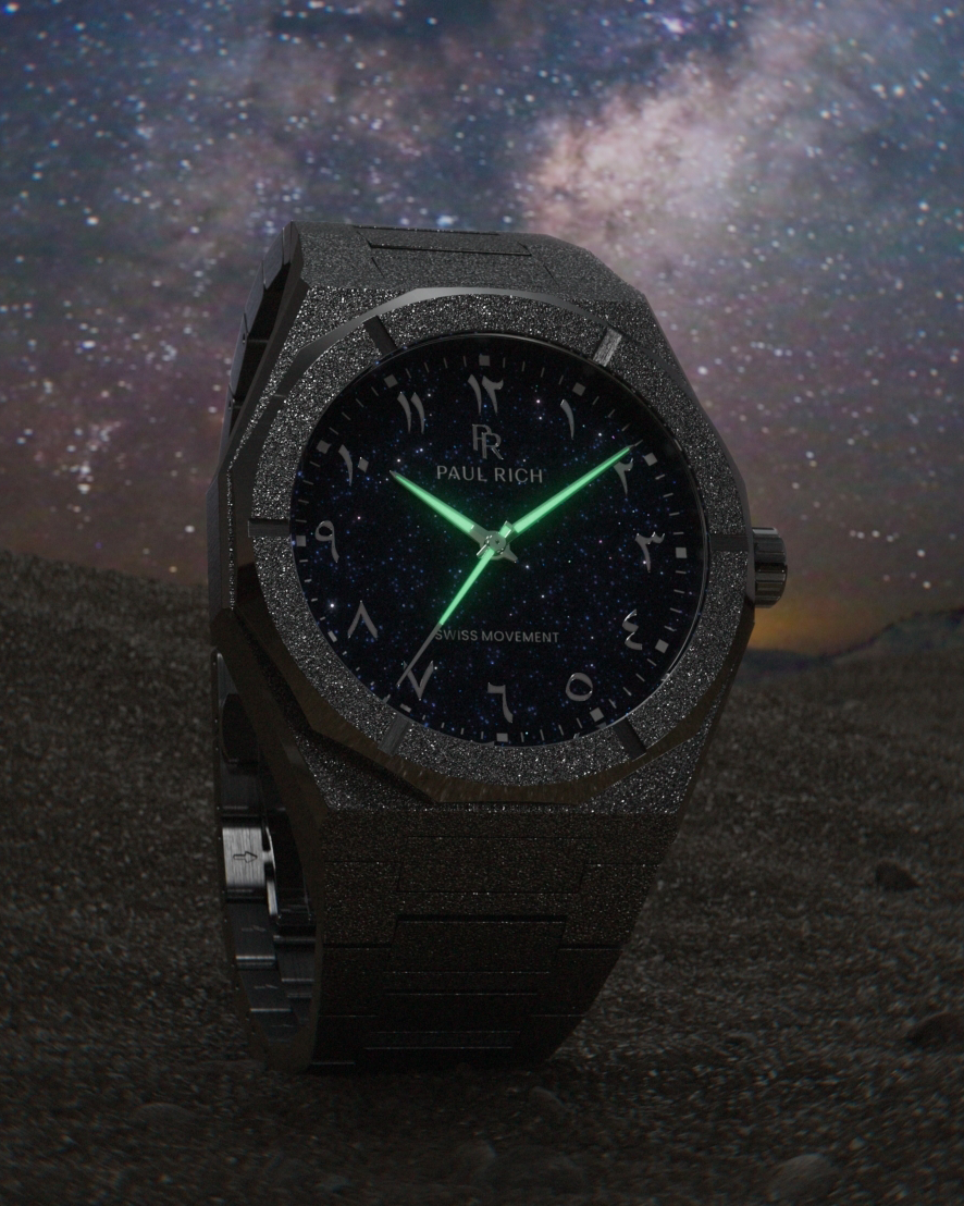 Frosted black watch with blue aventurine dial, Arabic numerals, and luminous green hands, set against a star-filled night sky over dark terrain.