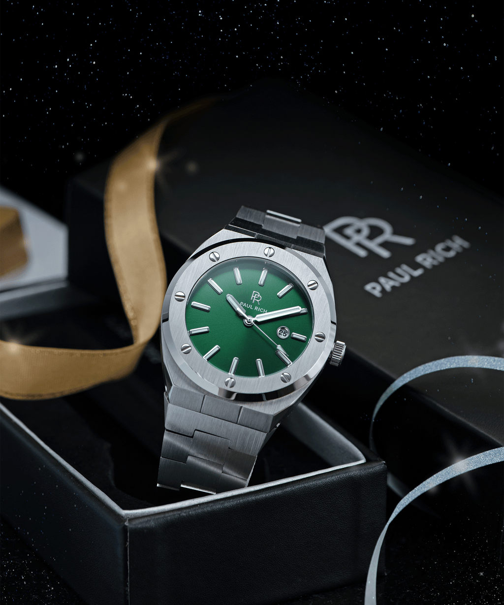 Paul Rich brushed silver watch with green dial and date window, displayed in open gift box with gold ribbon and starry night background.