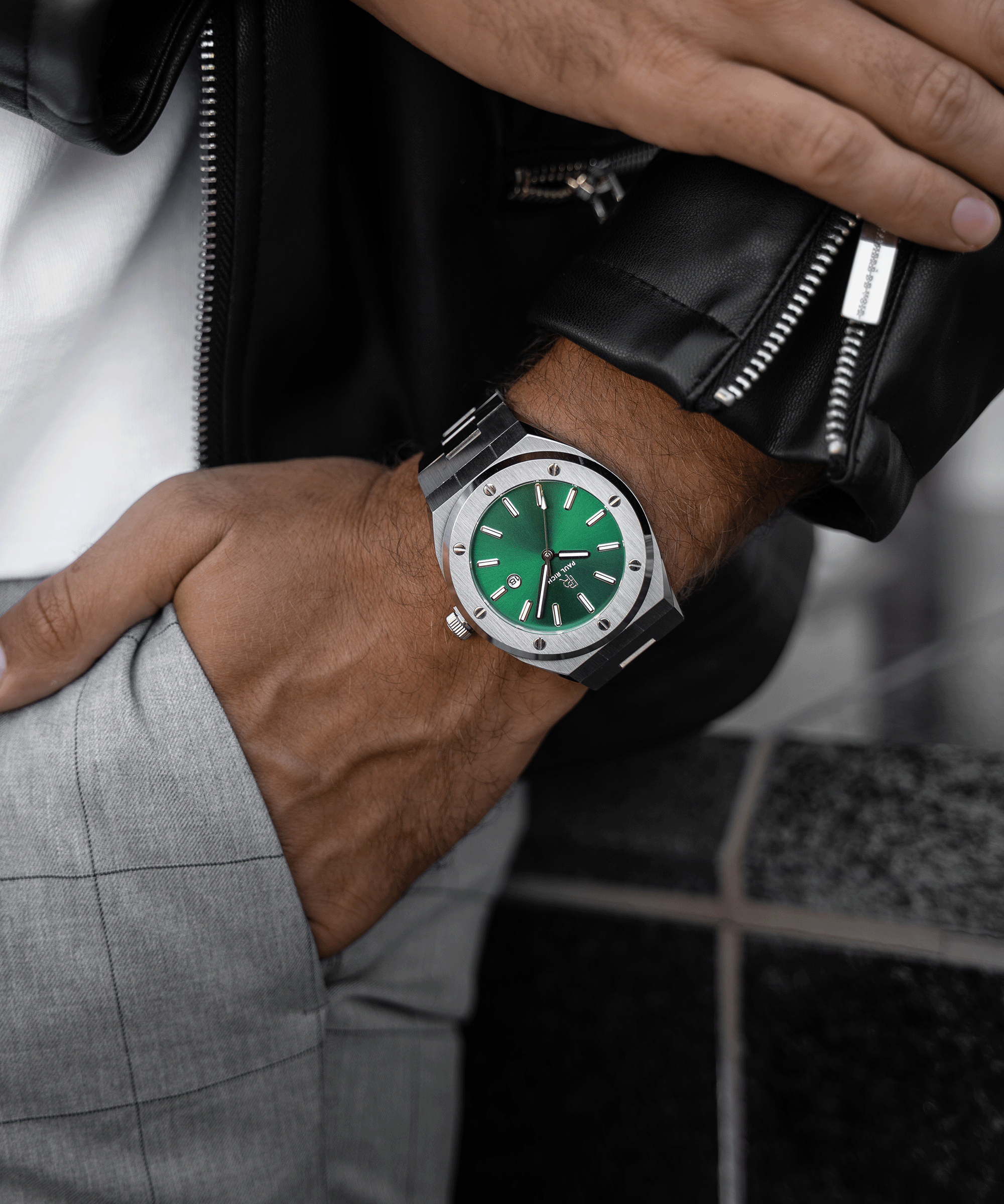 Paul Rich brushed silver watch with green dial and date window, worn with black leather jacket and gray plaid pants, hand in pocket.