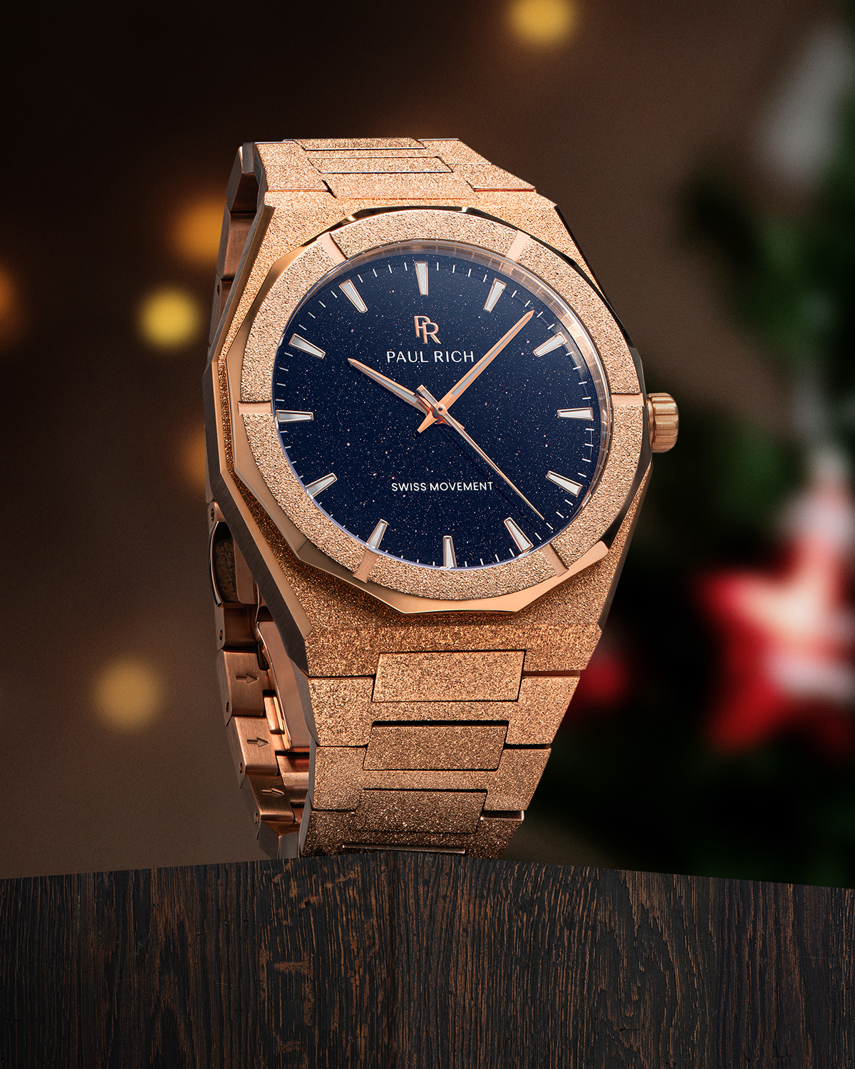 Rose gold Paul Rich watch with blue aventurine dial on wood surface, warm lights and blurred Christmas decor in the background.